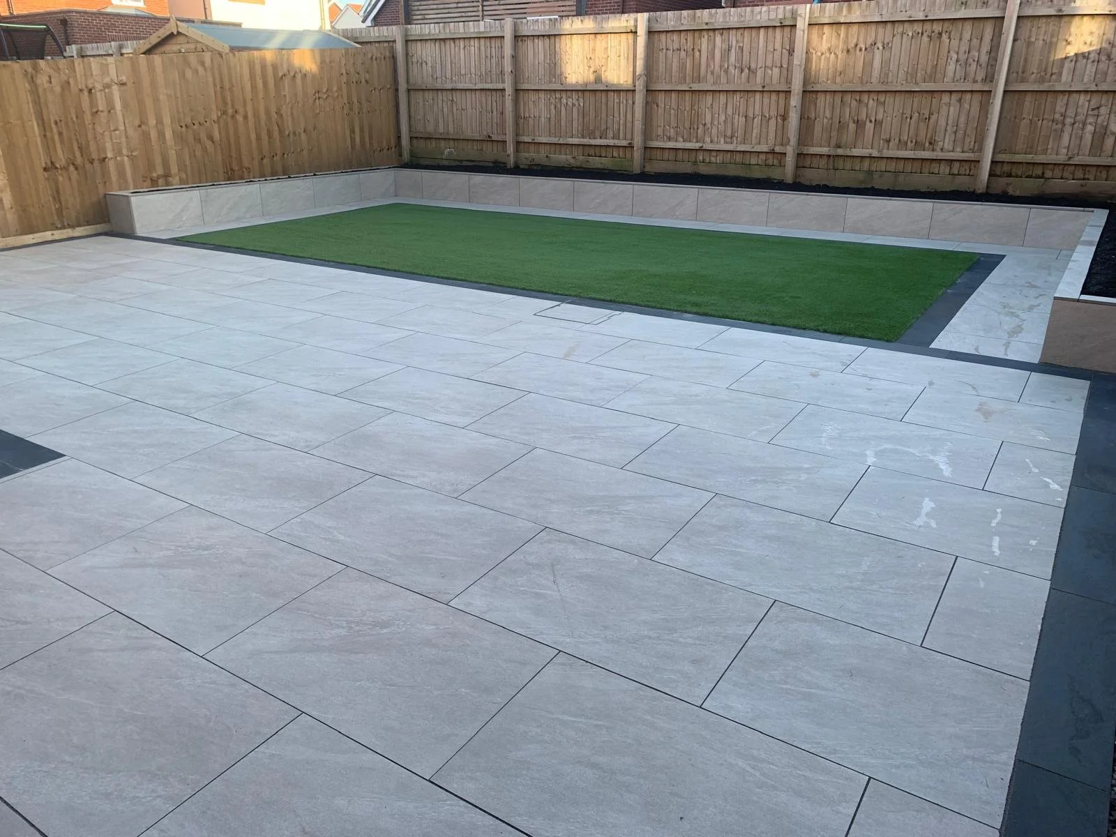 Backyard with a patio made of large light gray tiles, a rectangular grassy area, and a raised bed area with a concrete border, enclosed by a wooden fence.