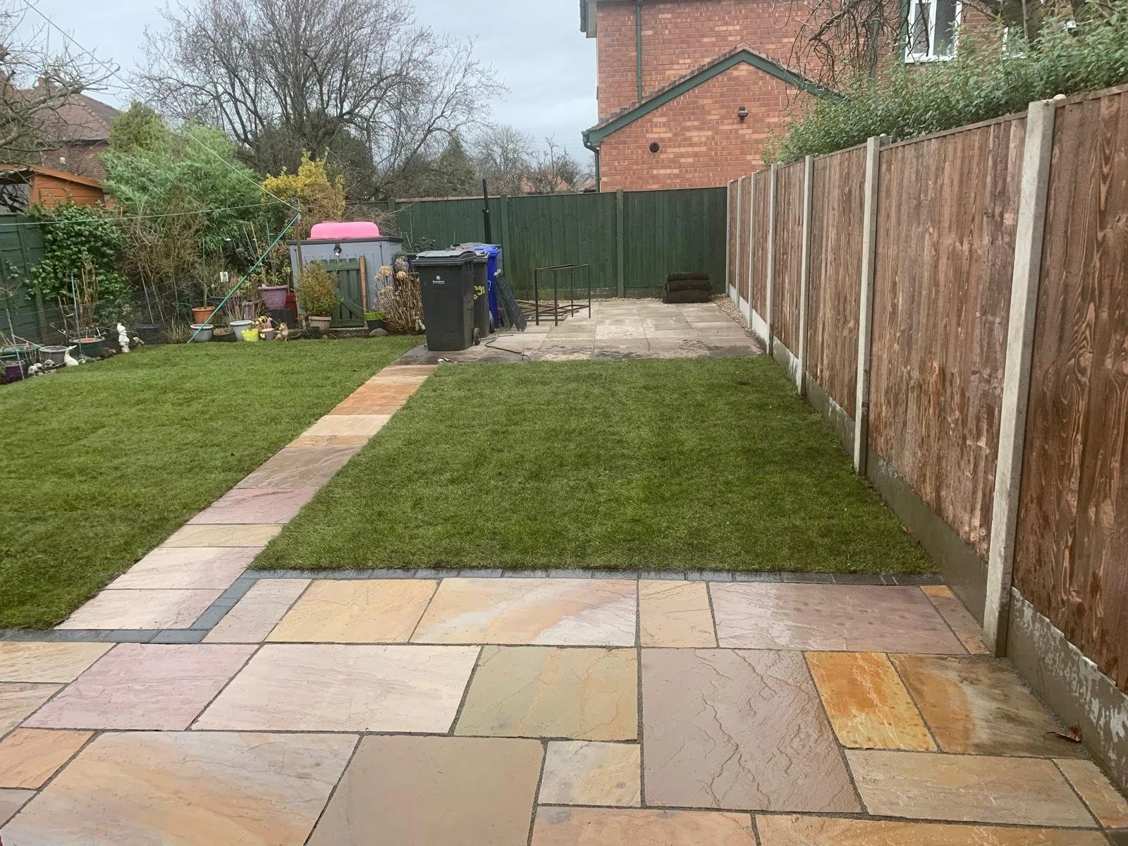 A backyard with a paved patio, a grassy lawn, and a wooden fence. There are plants and garden decorations on one side, waste bins near a shed, and a cloudy sky above.