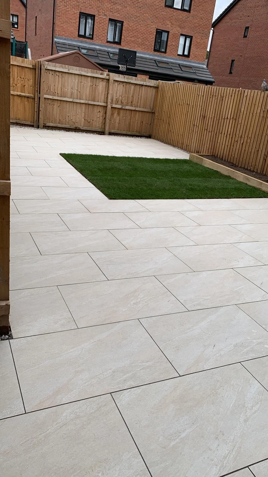 Backyard with light-colored tiled patio, small patch of grass, wooden fence, and neighboring brick buildings.