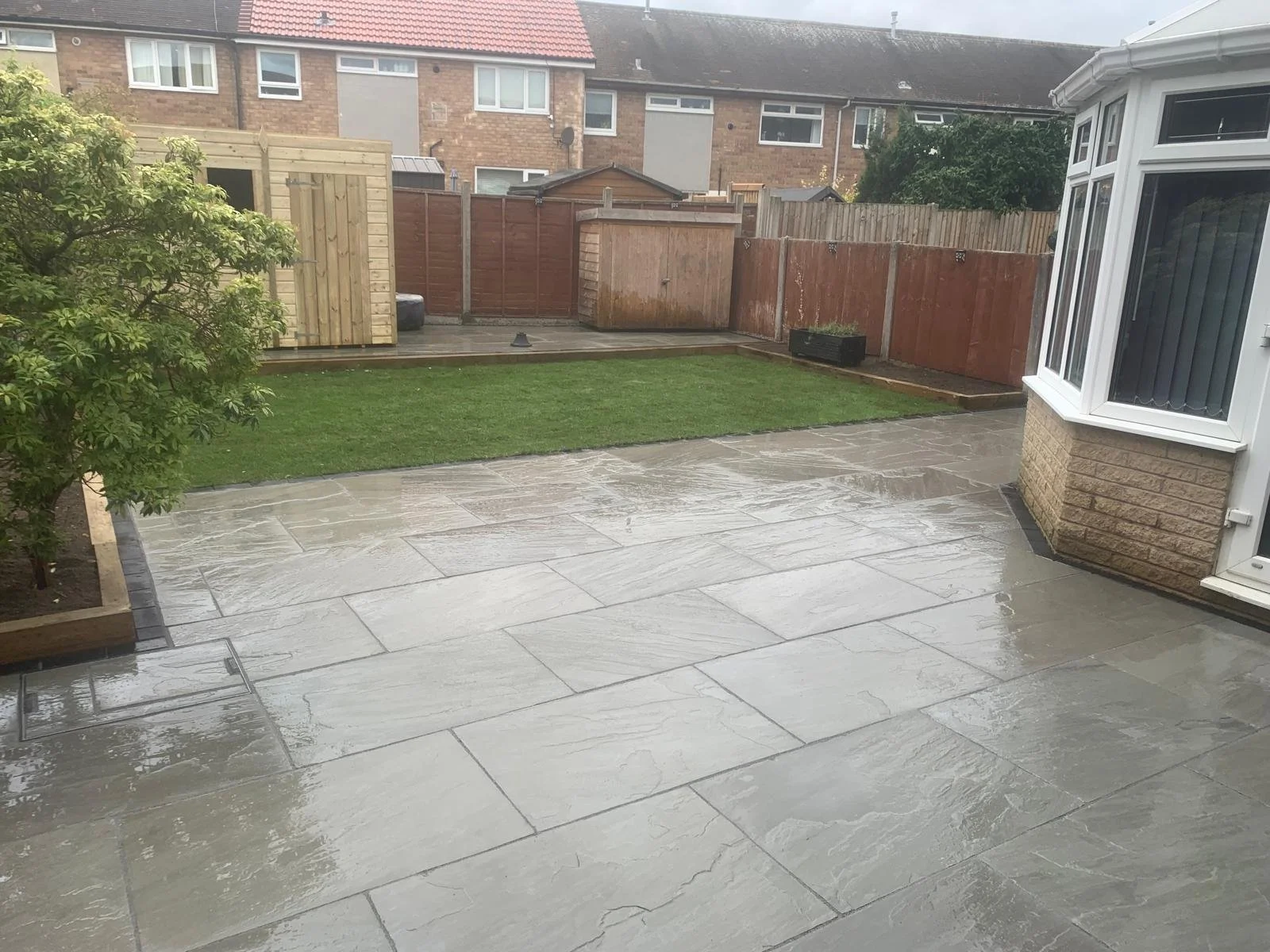 A backyard with wet, shiny stone patio, small grassy area, some trees, wooden fence, garden shed, and adjacent houses in the background.