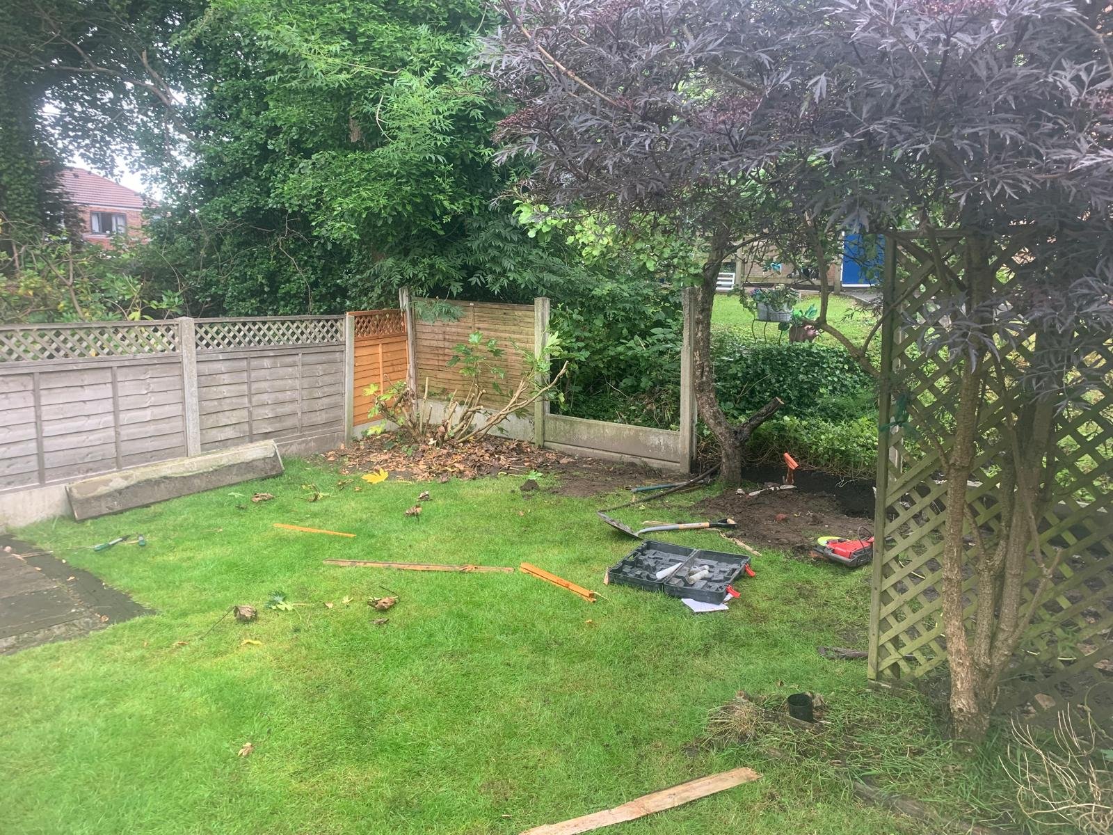 A backyard with a green lawn, a wooden fence partially in disrepair, and several tools and materials scattered on the ground, indicating ongoing gardening or fence repair work. There are trees and shrubbery around the area.