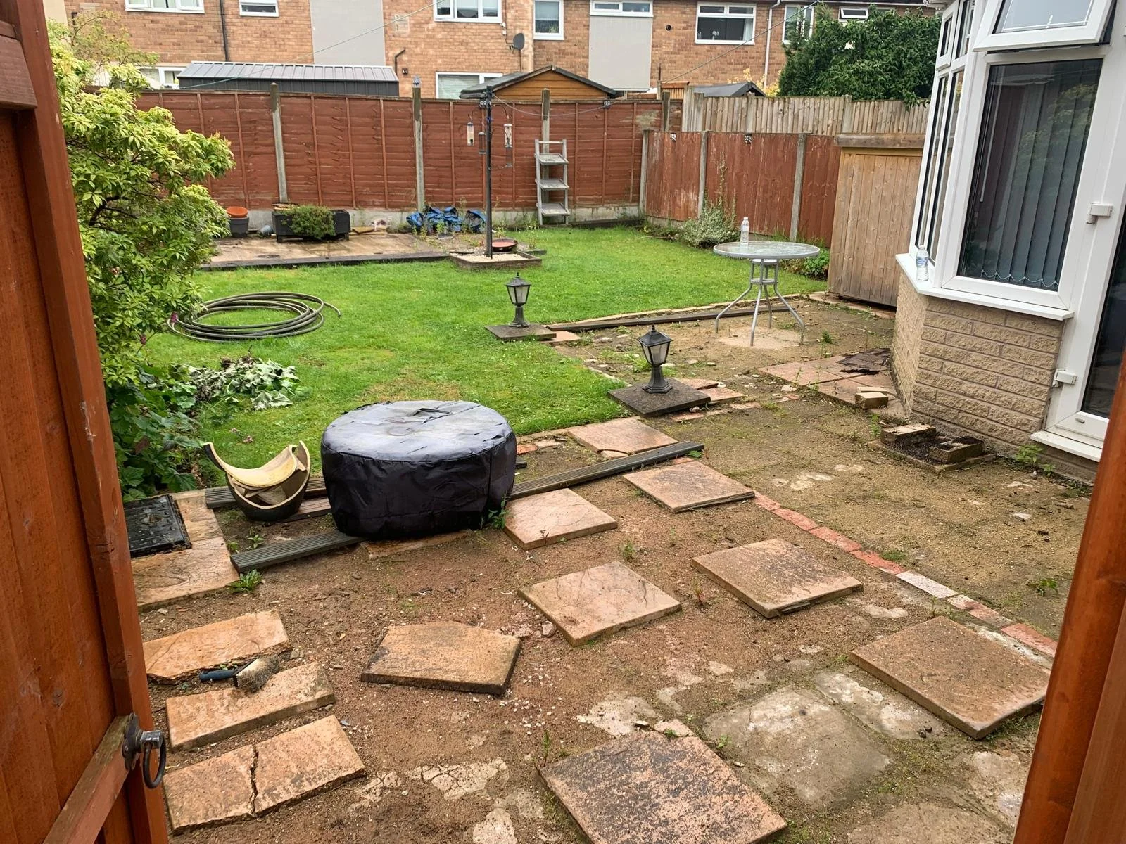View of a backyard with a green lawn, a paved area with stepping stones, garden lanterns, a table, a clothesline, a hose, and a shed, all enclosed by a wooden fence.