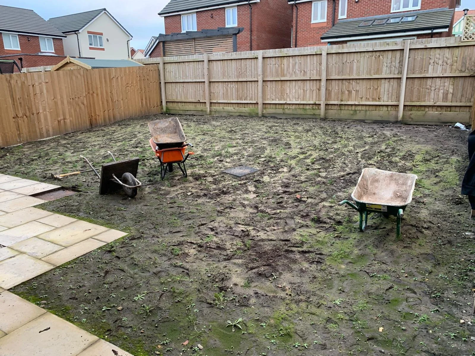 Bare backyard with dirt and some small weeds, enclosed by wooden fences, with two partially filled wheelbarrows, a garden trowel, and a few paving stones on a paved patio area.