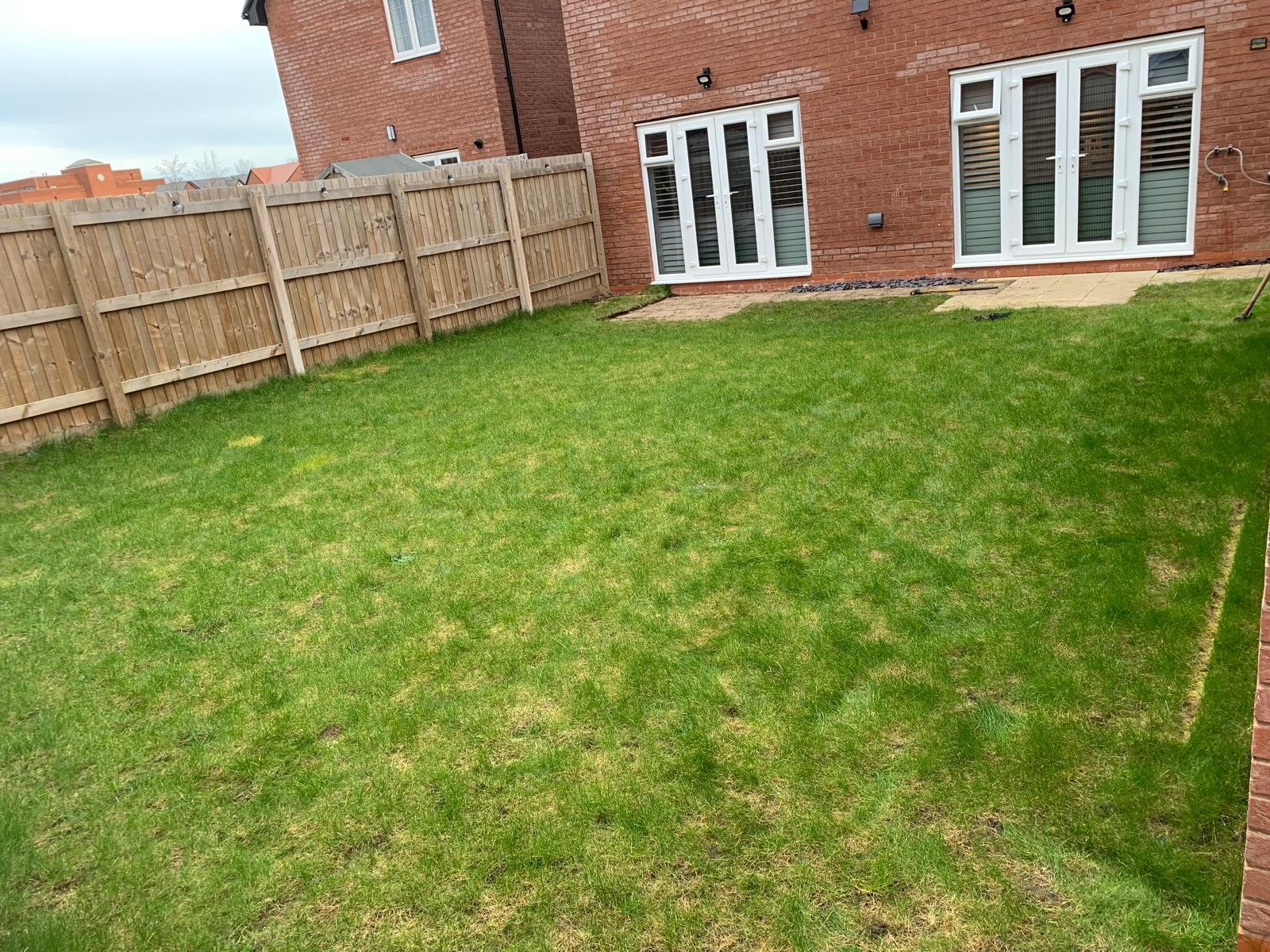 A backyard with green grass, a wooden fence, and a brick house with white French doors and a window, some paving stones and a small patch of dirt near the house.