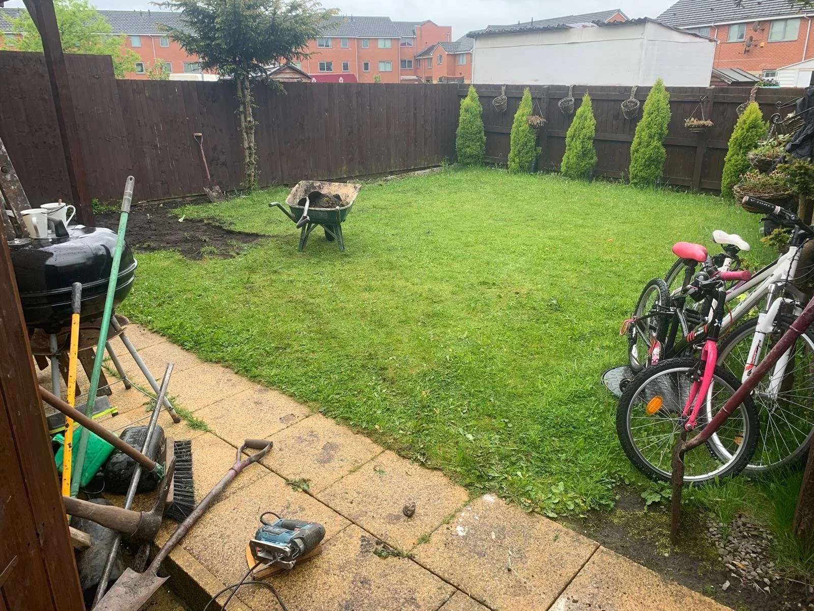 A backyard with a grassy lawn, a wooden fence, and a paved patio area. There are two bicycles leaning against the fence on the right, a wheelbarrow on the grass, and gardening tools and supplies on the patio in the foreground.