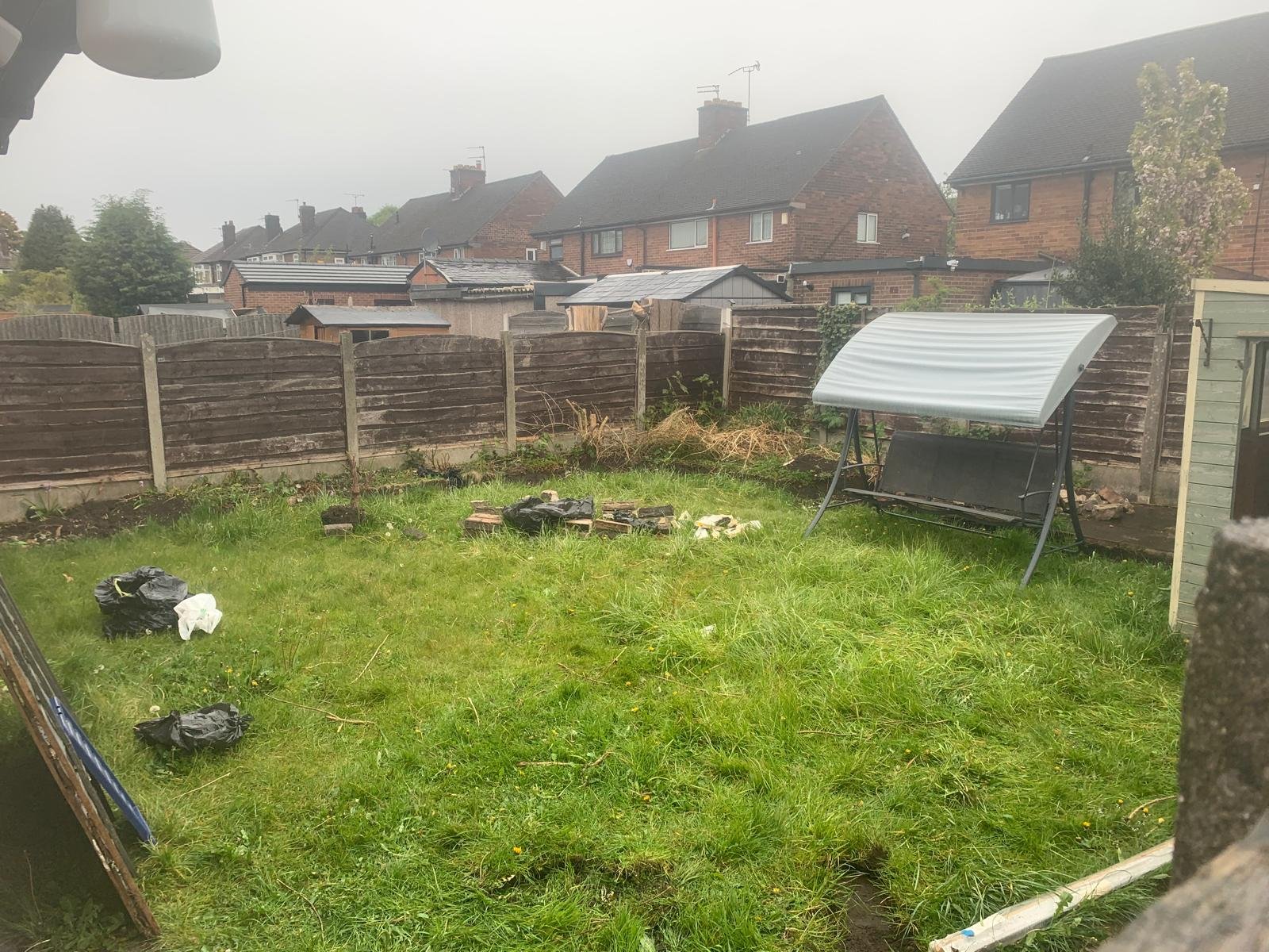 Backyard with a green lawn, trash bags, bricks, a shed, and a wooden fence, with neighboring houses visible in the background under overcast weather.