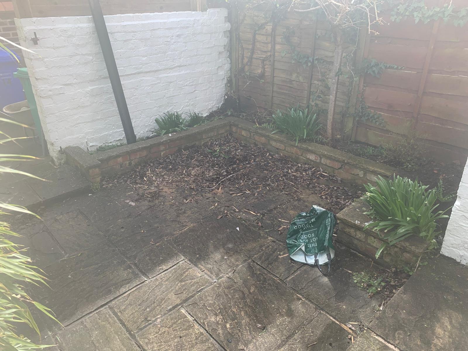 Empty garden bed with some plants and fallen leaves, surrounded by brick edging, next to a paved patio area and a white brick wall.