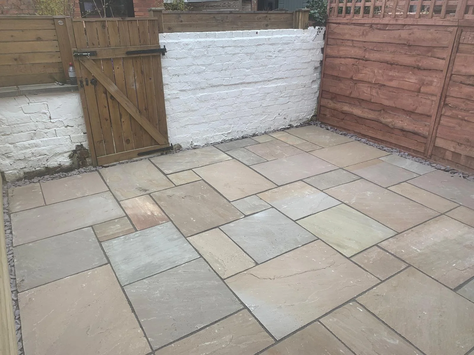A backyard patio with large beige and gray stone tiles surrounded by a wooden fence. There is a small wooden gate on the left side and a white painted brick wall on the back, with a section of unpainted brick on the left. Rocks are lining the area be