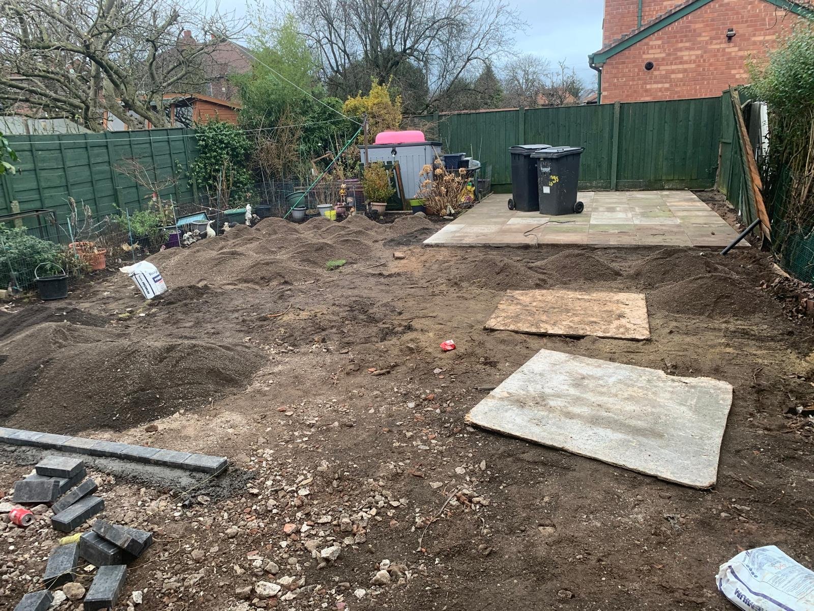 A backyard with dirt and construction materials, preparing for paving or landscaping, with a fenced area, potted plants, and trash bins in the background.