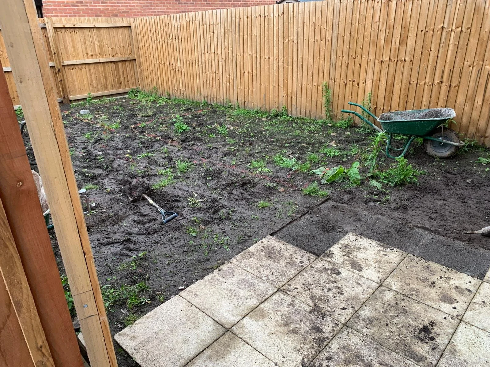 A backyard garden area with dark soil, small green plants, and weeds, enclosed by a wooden fence, with a wheelbarrow and garden tools. Paved stone stepping stones are at the foreground.