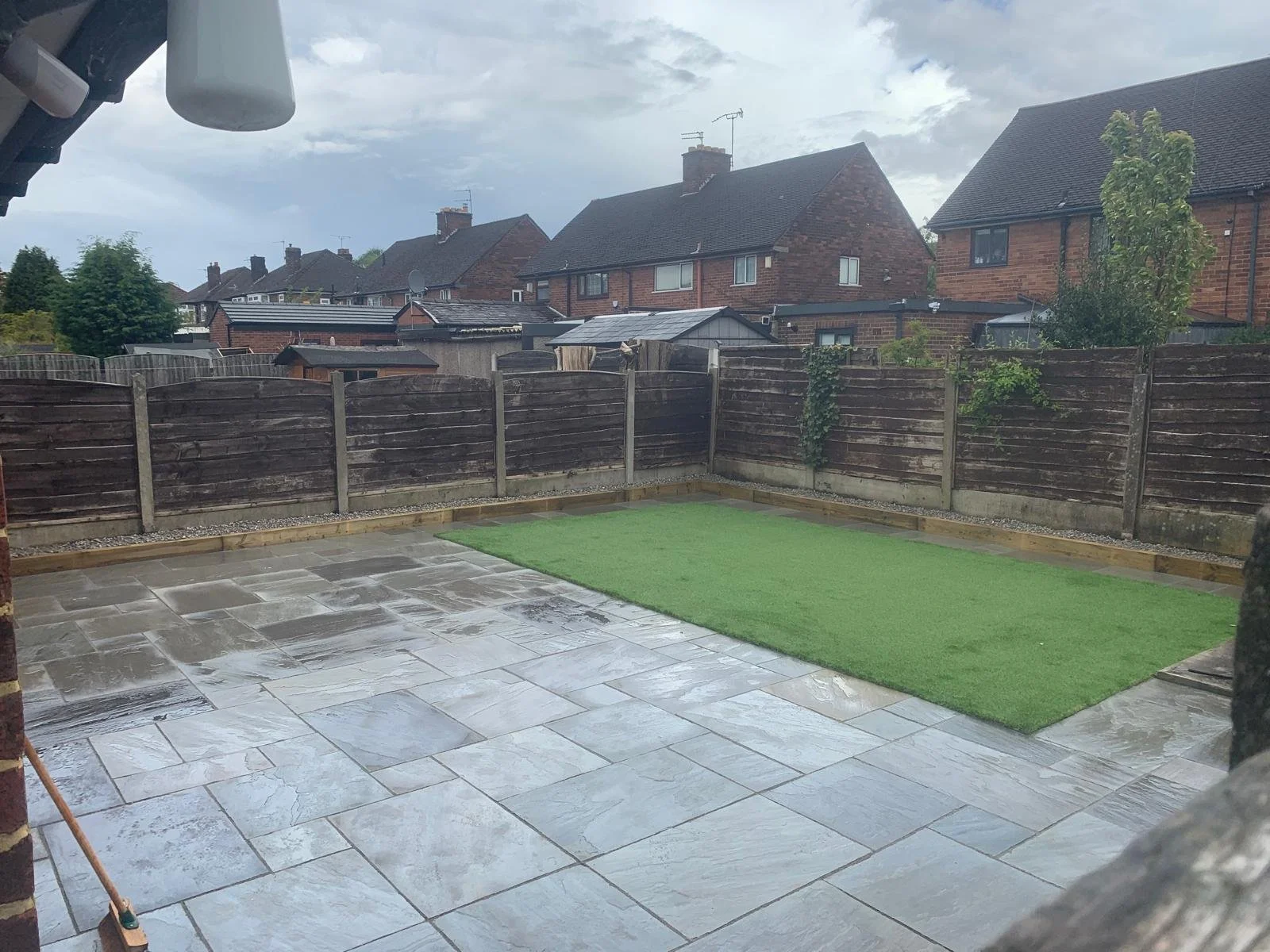 A backyard patio with wet stone tiles, a small patch of artificial grass, and a weathered wooden fence. Overcast sky with dark clouds in the background.