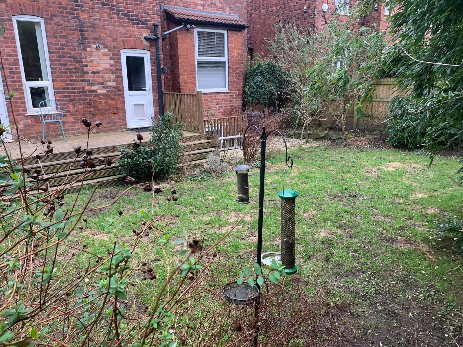 A backyard garden with a grass lawn, bushes, small trees, two bird feeders on stands, a bird bath, a small deck with a chair, and brick house walls with windows and a door.