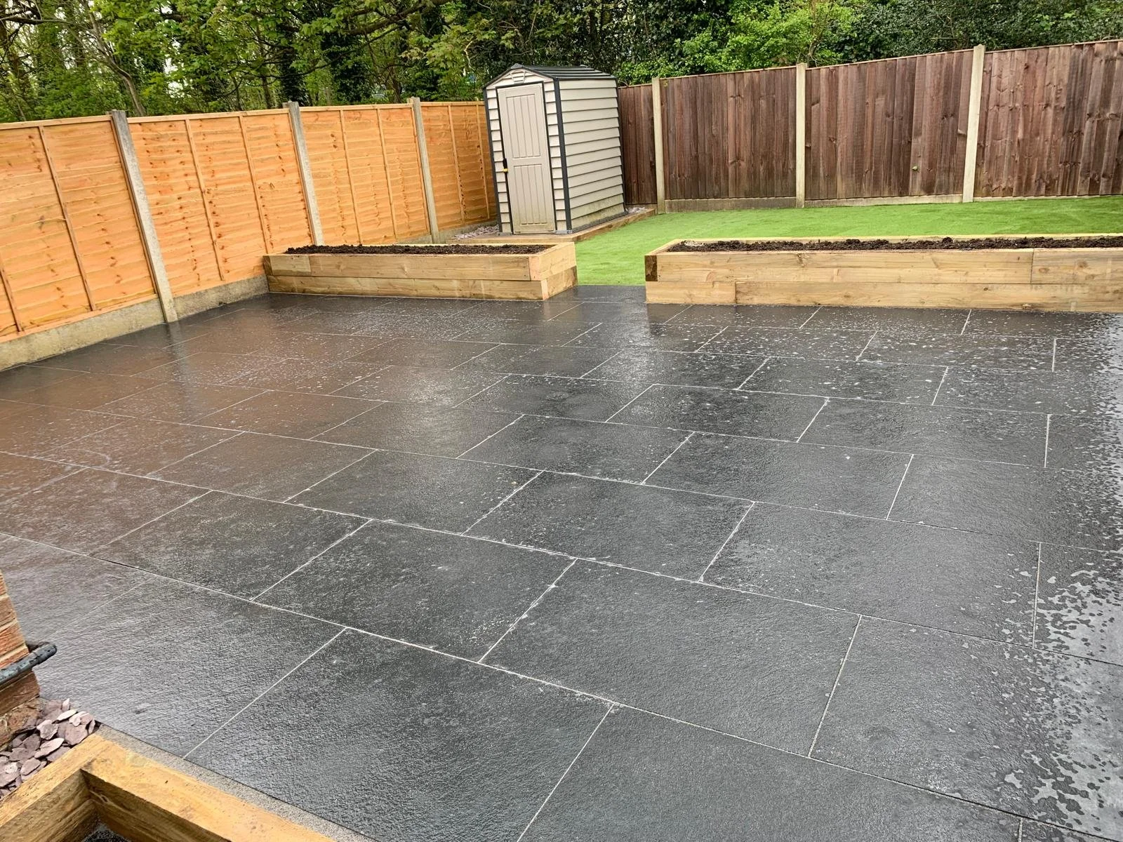 Backyard area with wet black stone patio, wooden planter boxes filled with soil, green grass, a shed, and a wooden fence.