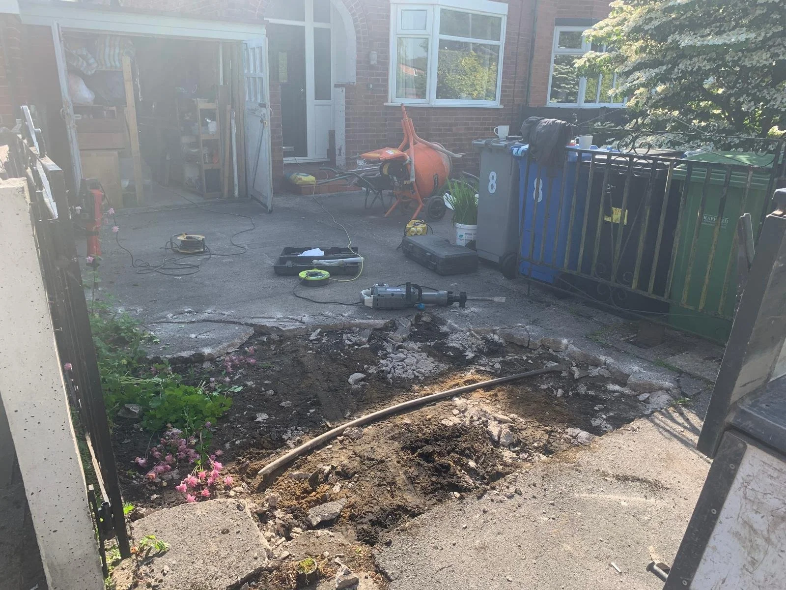 Front yard of a house under construction or repair with a hole in the ground, construction tools, and equipment including a drill, extension cord, and work lights. The driveway is partially dug up, and there are flower beds and garden items around.