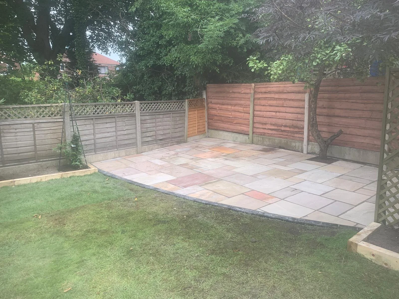 Fenced backyard with a tiled patio, grassy area, garden bed, tree, and surrounding wooden fences.