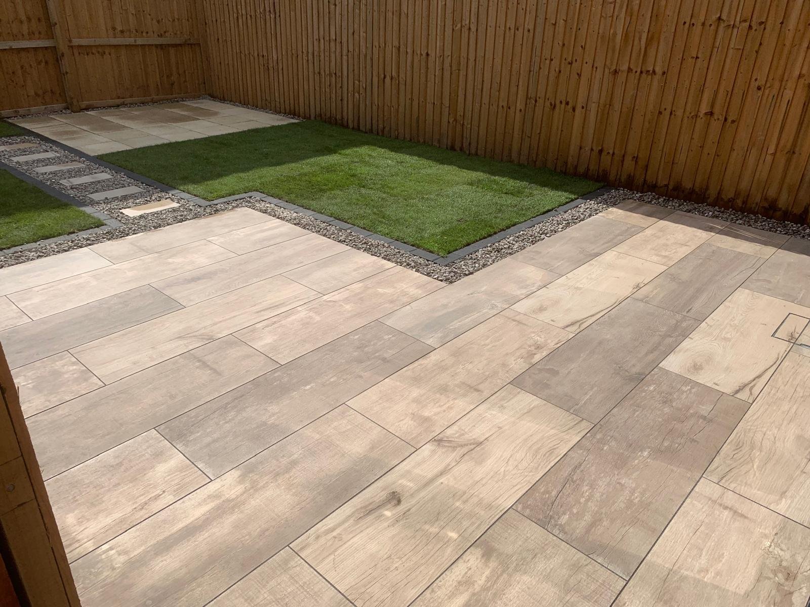 Backyard with large beige paver patio, small patches of artificial grass, and a wooden fence.
