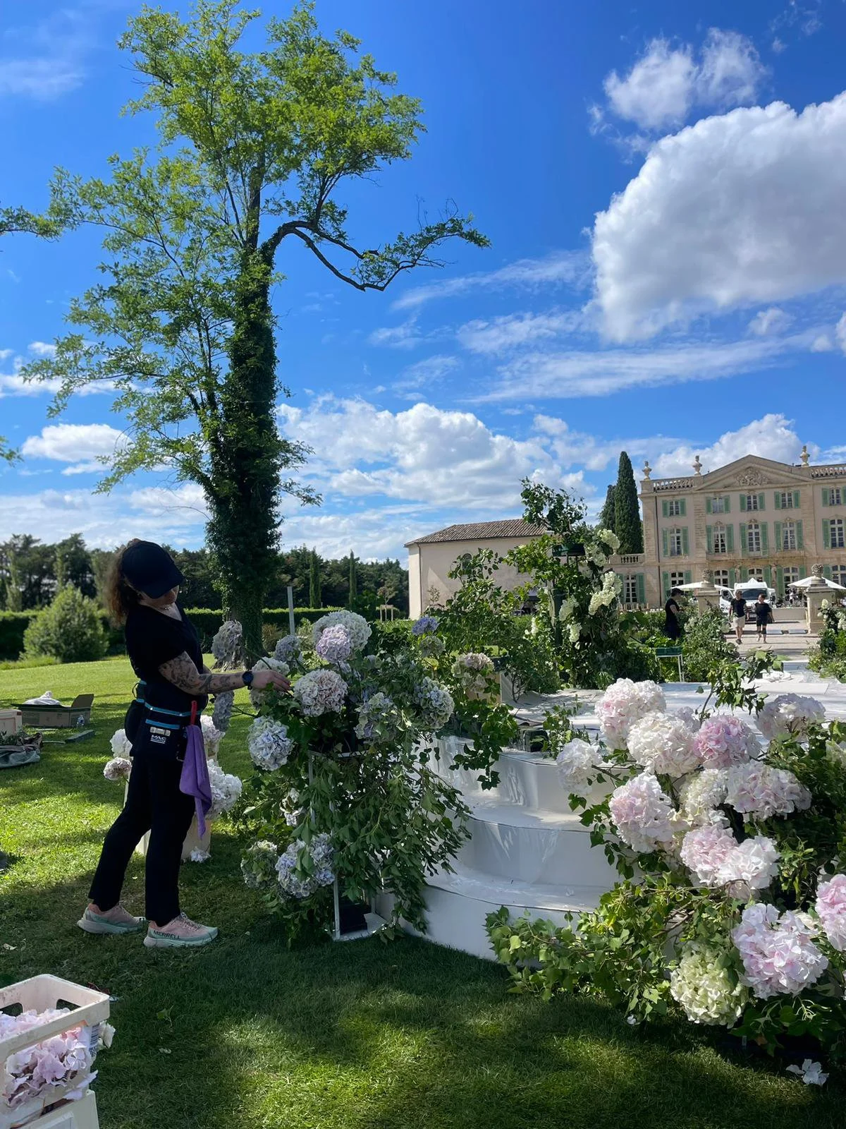 Une femme arrange des bouquets de fleurs dans un jardin en plein air, avec un bâtiment élégant en arrière-plan, sous un ciel bleu parsemé de nuages.