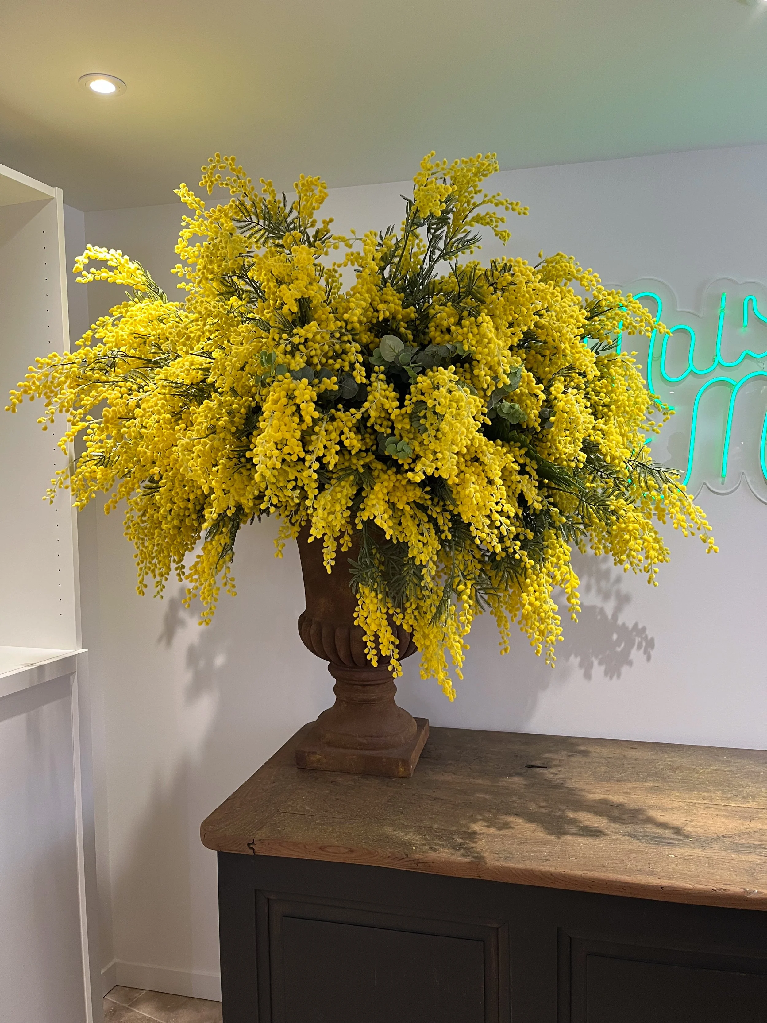 Grand bouquet de fleurs jaunes dans un vase en terre cuite sur une table en bois, avec un mur blanc en fond et un panneau lumineux vert à droite.