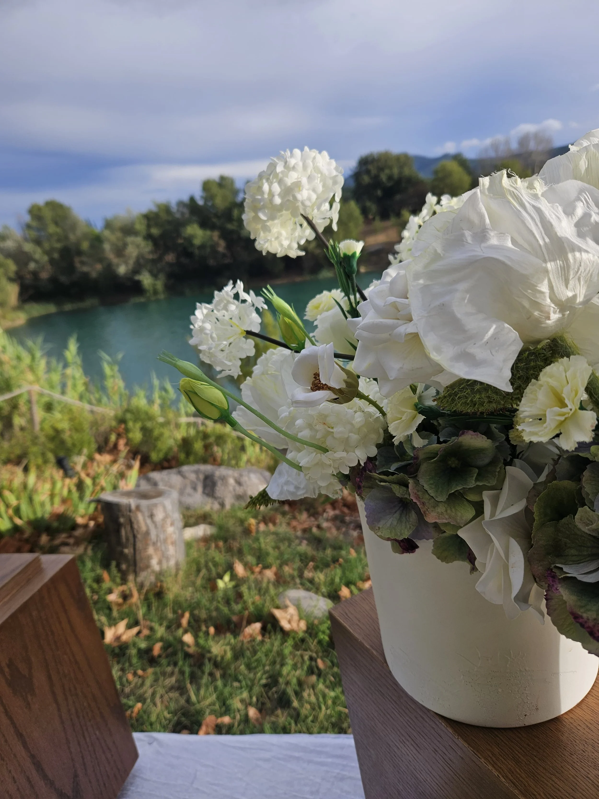 Un bouquet de fleurs blanches dans un vase blanc sur une table en bois, avec un paysage extérieur comprenant un lac, des arbres et un ciel nuageux en arrière-plan.