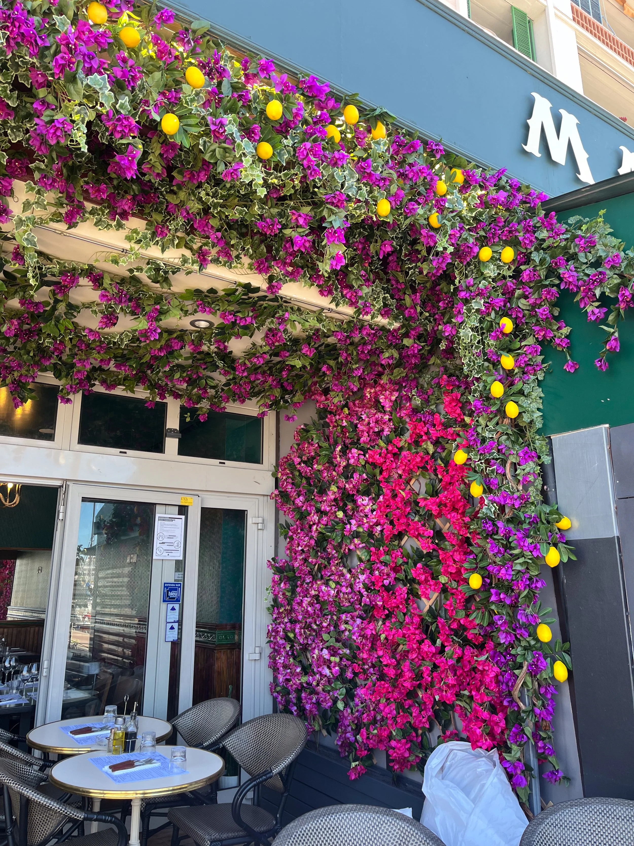 Terrasse de restaurant avec tables et chaises, décorée de fleurs de bougainvillier violettes et quelques citrons jaunes suspendus.
