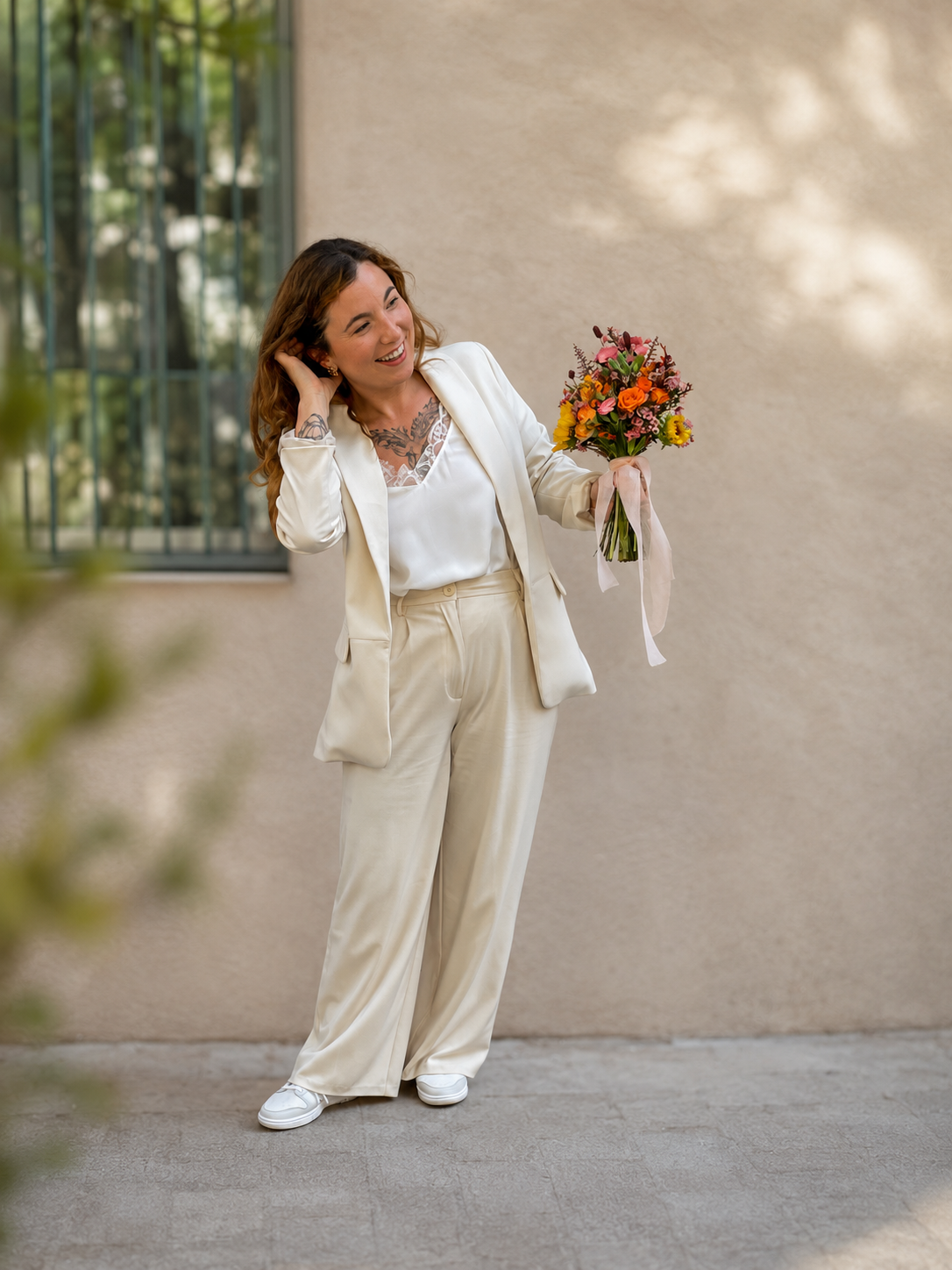 Une femme souriante habillée en blanc, tenant un bouquet de fleurs, se tenant devant un mur beige avec une fenêtre en arrière-plan.