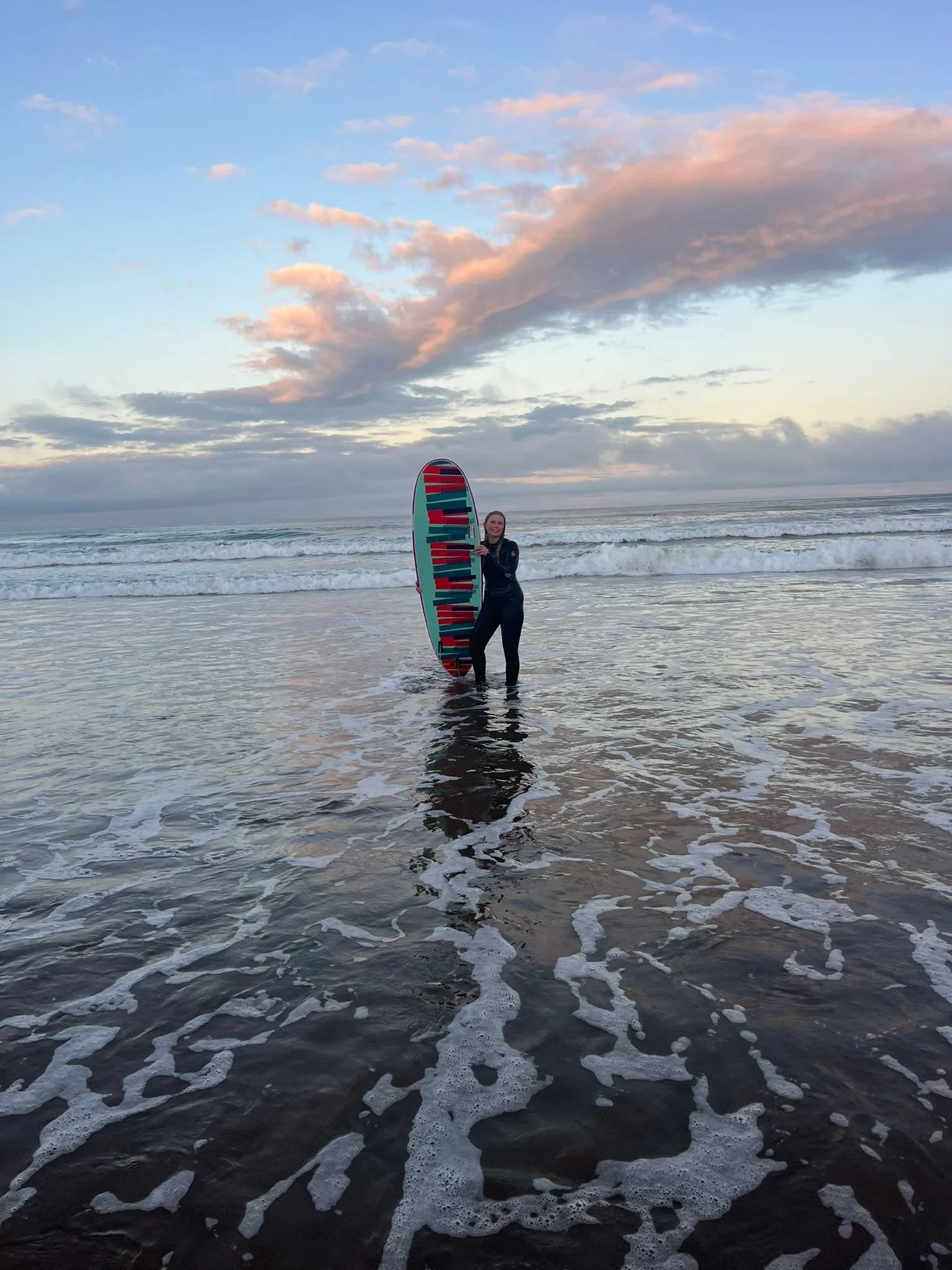 A woman in black wetsuit holding a colorful paddleboard standing in shallow ocean water at sunset, with waves and a sky filled with clouds and soft pink and blue hues.