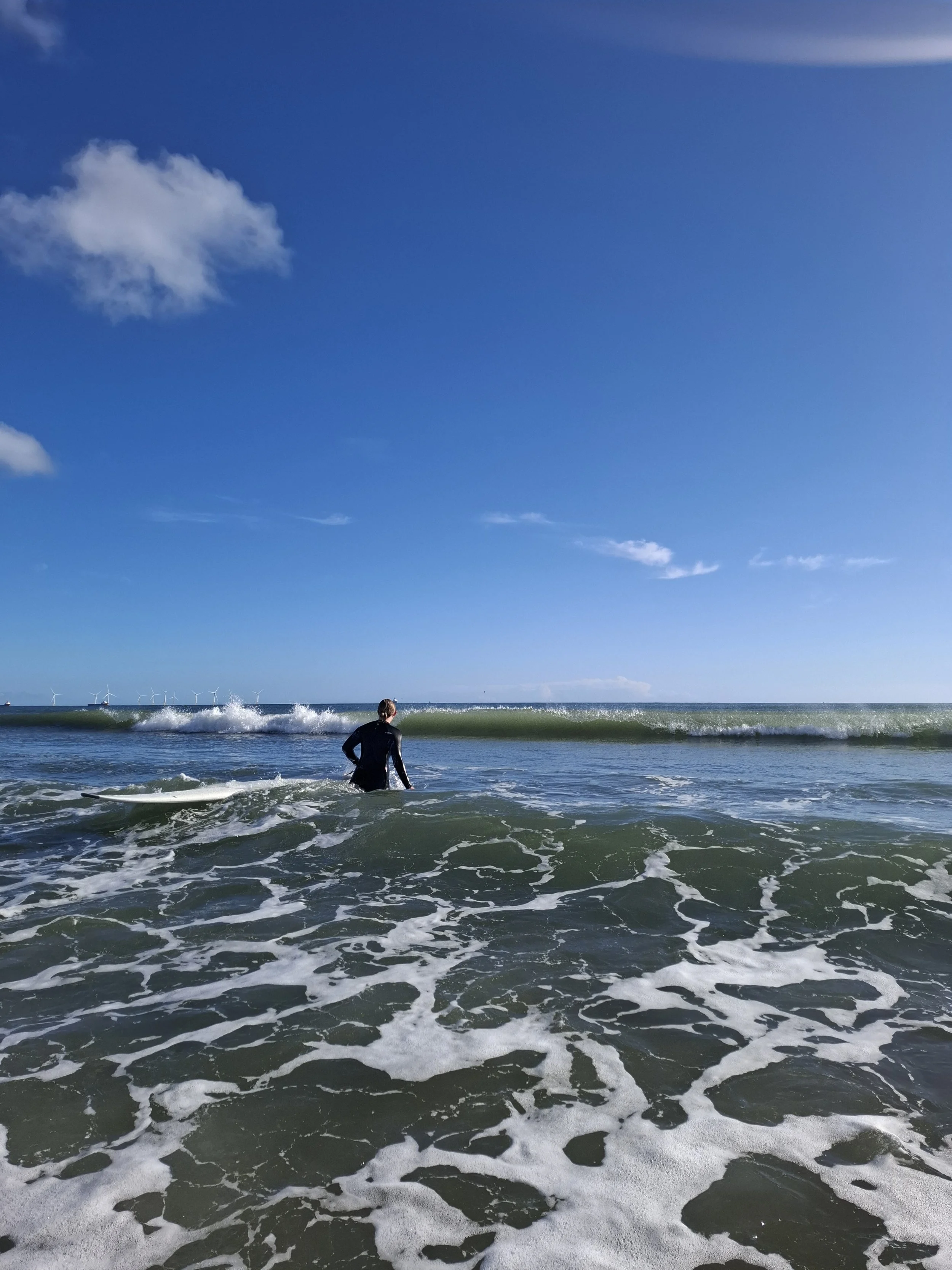 Person standing on the shore near the ocean, with waves crashing at their feet and wind turbines in the distance under a partly cloudy blue sky.