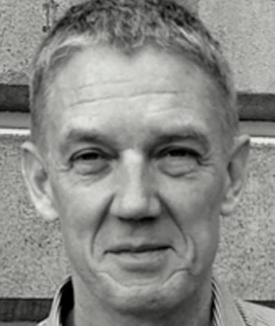 Close-up black and white photo of a middle-aged man with short, light hair, looking slightly to the side, standing in front of a concrete wall.