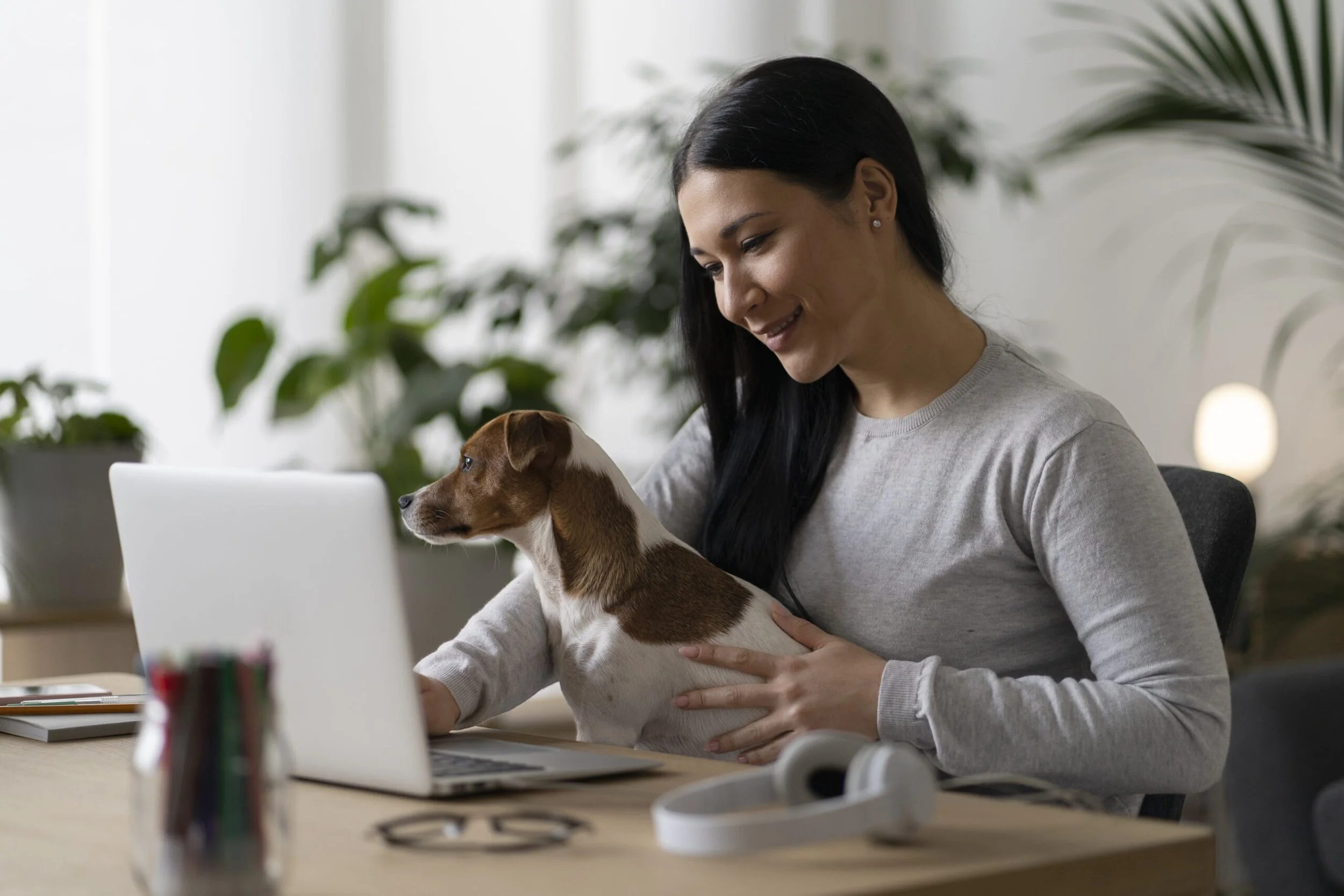 A woman with long dark hair in a ponytail smiling and holding a small brown and white puppy while working on a laptop in a bright room with green plants in the background.