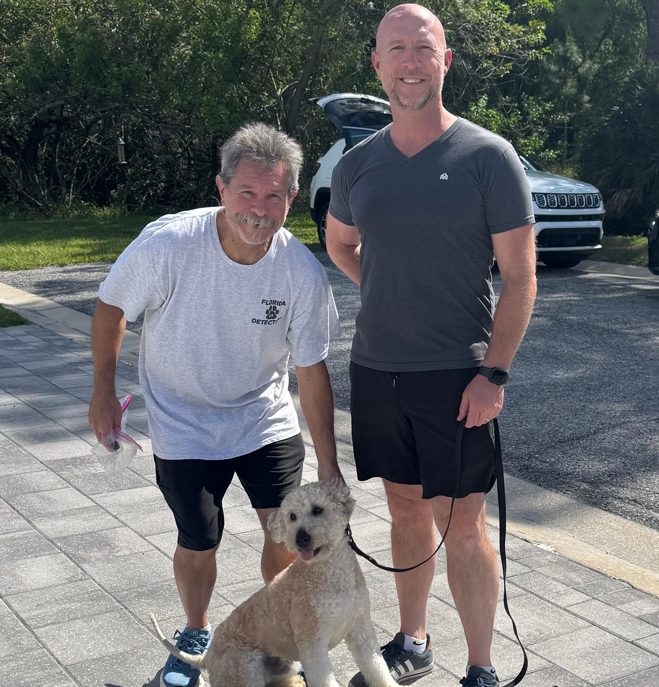 Two men and a dog standing on a sidewalk with trees and parked cars in the background. One man is bending slightly, wearing a white T-shirt with a skull design, and the other man is standing upright, wearing a dark gray T-shirt. The dog is a fluffy, light-colored breed with a leash, sitting and looking at the camera.
