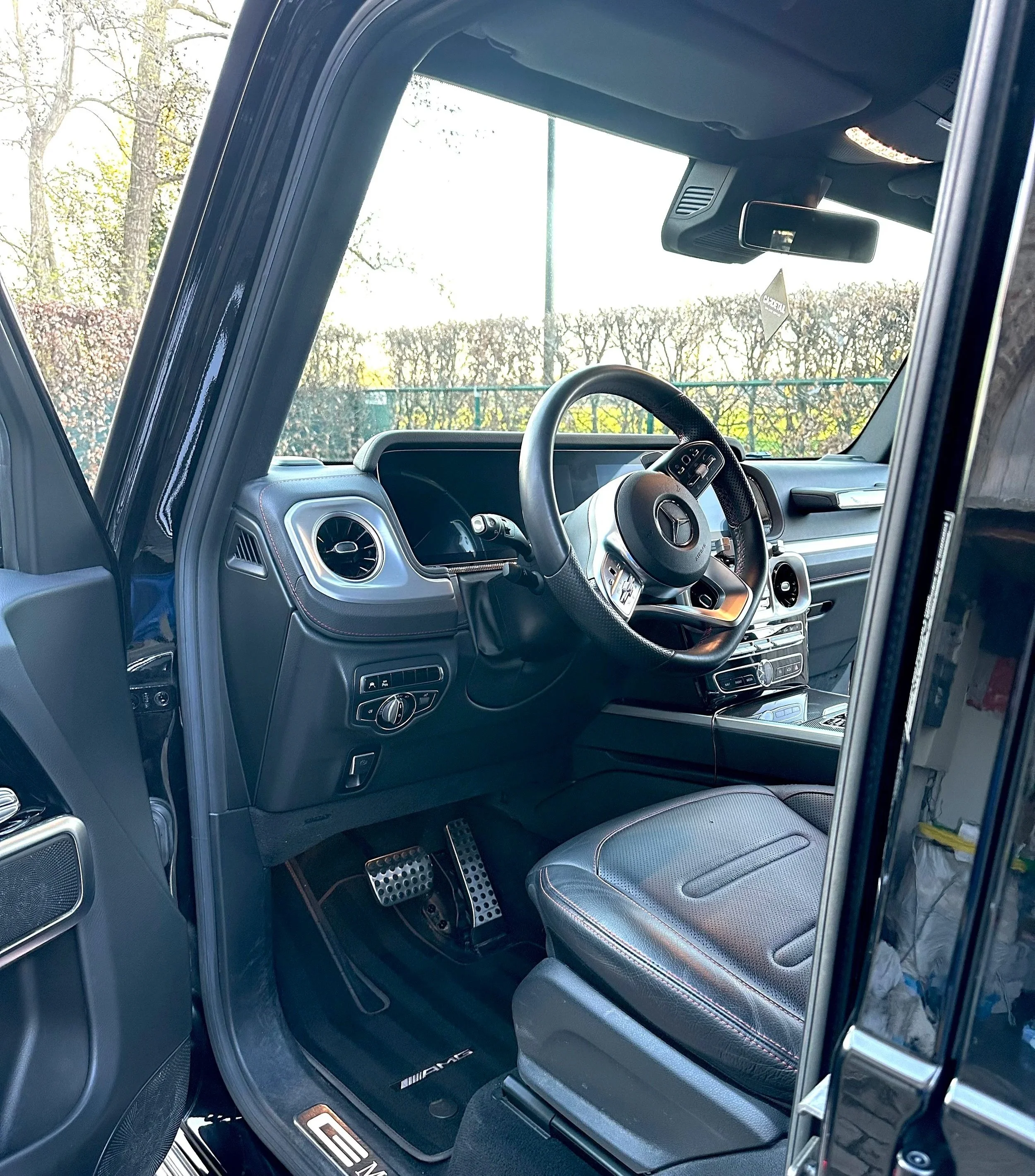 The interior of a Mercedes-Benz vehicle, showing the dashboard, steering wheel, driver's seat, and part of the passenger seat. The dashboard features air vents, controls, and a scenic view of trees through the windshield.