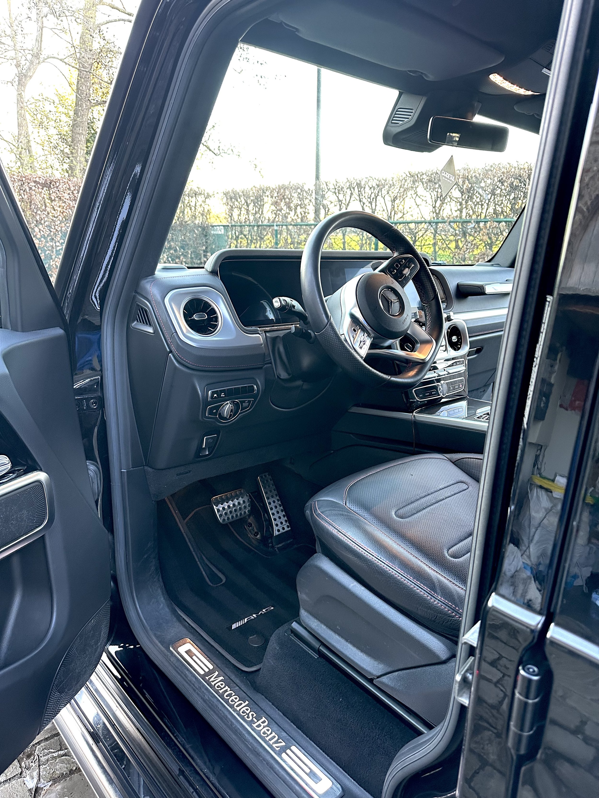 The interior of a Mercedes-Benz vehicle, showing the driver's seat, steering wheel, dashboard, and floor mats with Mercedes-Benz branding, with the driver's side door open.