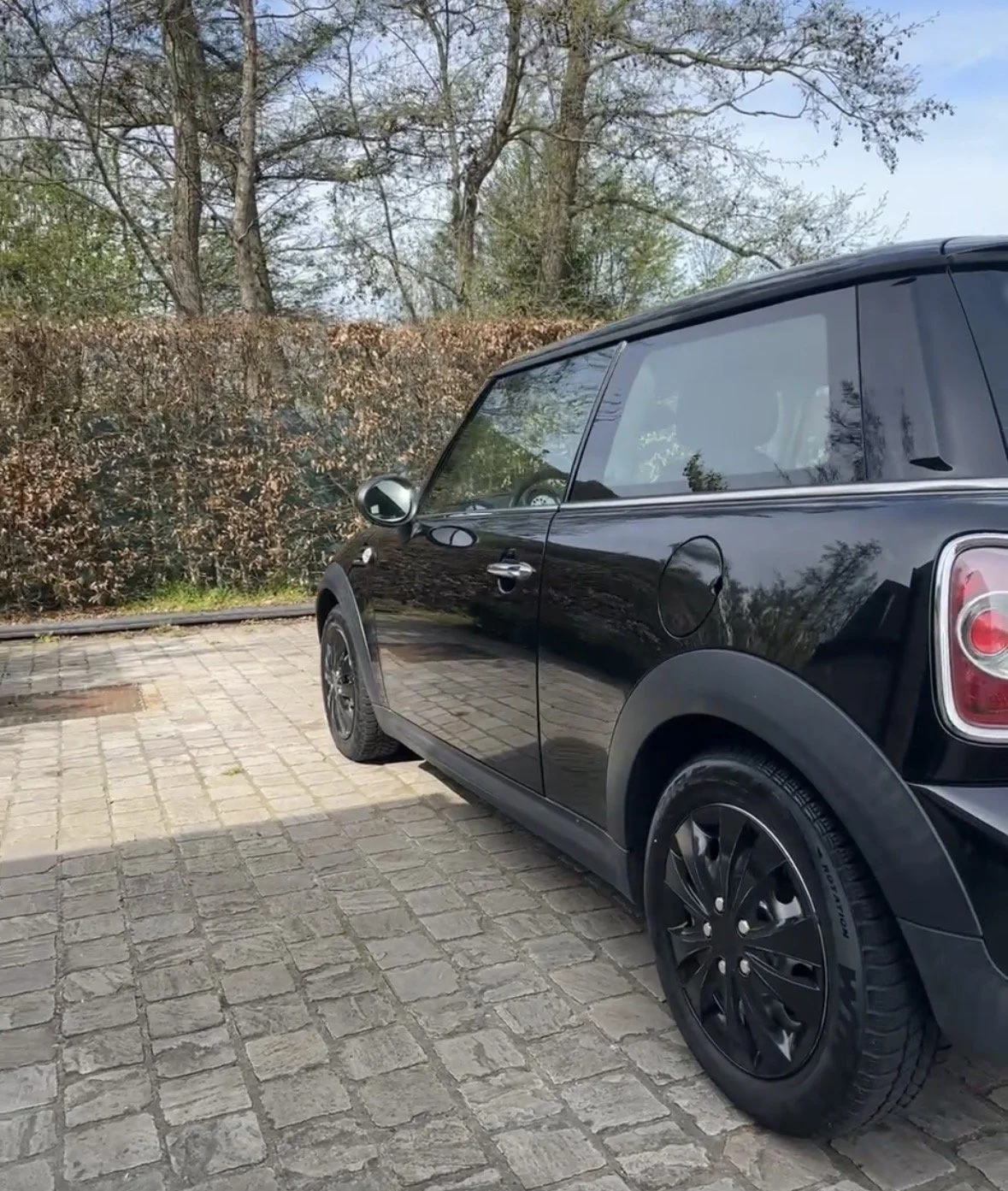 A black compact car parked on a cobblestone surface with a hedge and trees in the background.