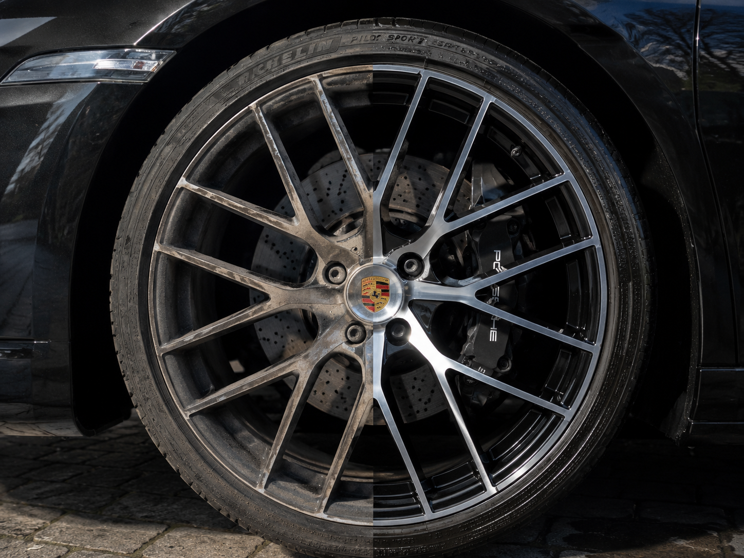 Close-up of a black car's front wheel with a silver multi-spoke rim and a Porsche emblem at the center, showing brake caliper and drilled brake disc behind the rim.