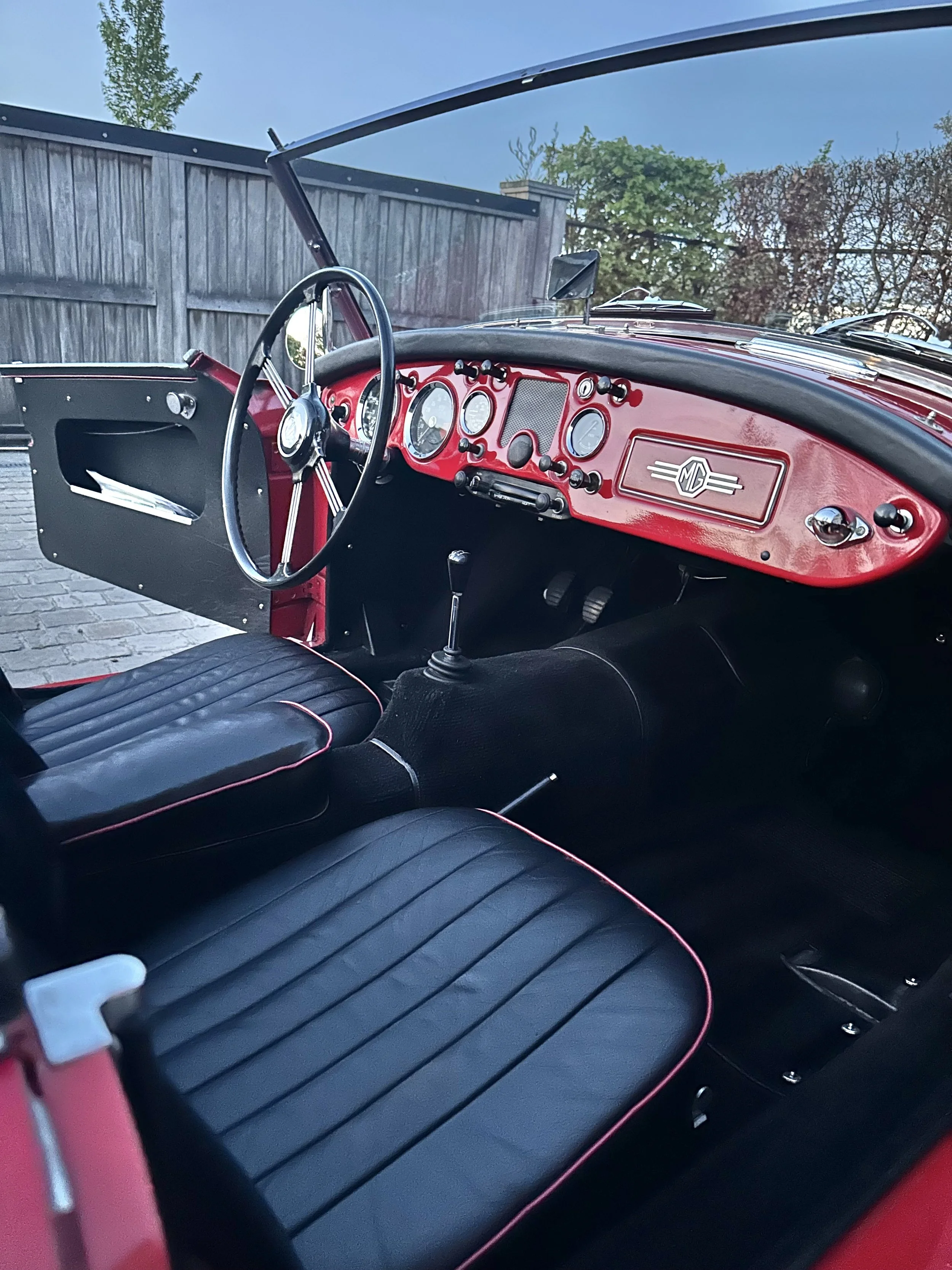 Interior of a vintage red MG convertible car with black seats and a dashboard with multiple gauges and controls,, with a wooden fence and trees in the background.
