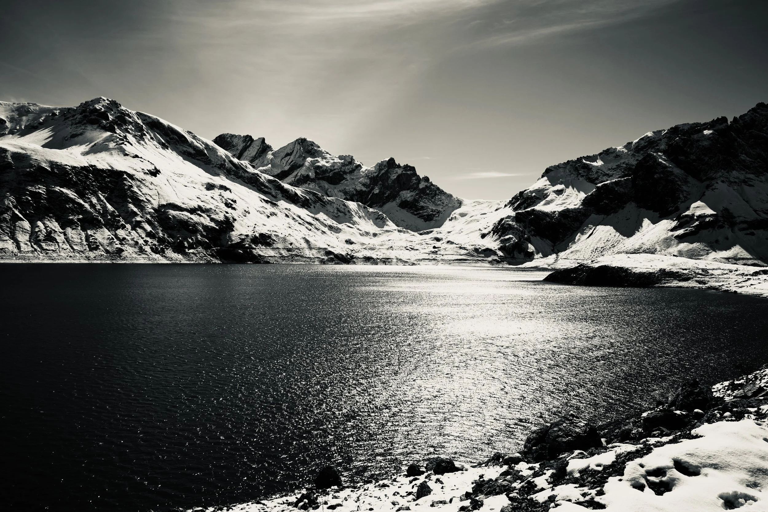 Schneebedeckte Berge und ruhiger See unter bewölktem Himmel in einer düsteren, monochromen Szenerie.
