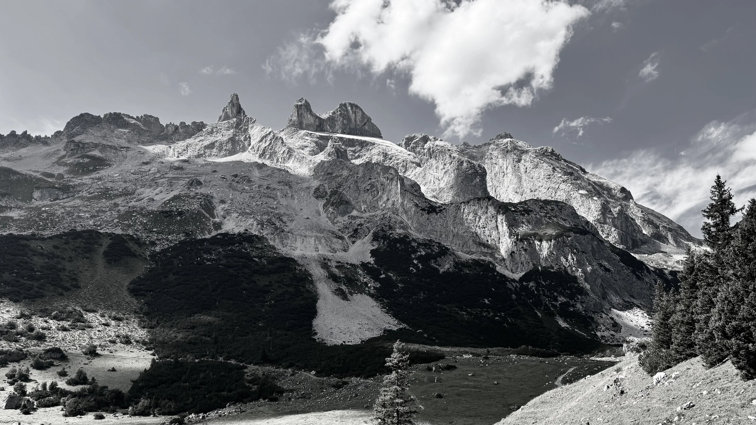 Berglandschaft mit hohen, schneebedeckten Gipfeln und dunklen Wäldern im Vordergrund. Wolken ziehen am Himmel vorbei.