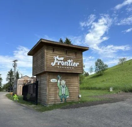 Petite structure en bois avec un panneau indiquant 'Frontier'. Sur le mur, il y a une peinture d'un personnage en costume traditionnel. Le ciel est en partie nuageux, et il y a une colline herbeuse à l'arrière.