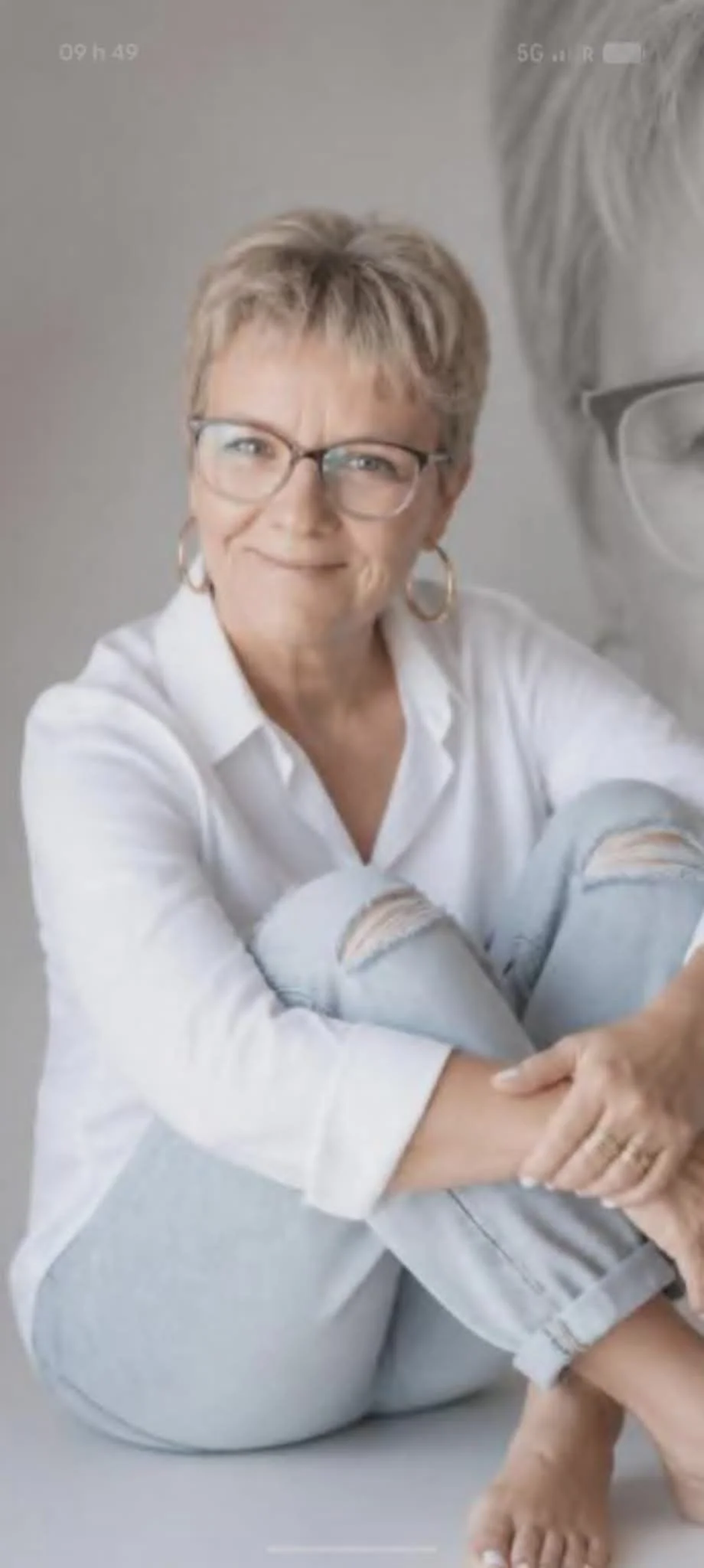 Une femme souriante, portant des lunettes et une chemise blanche, assise avec les jambes croisées, dans un style décontracté.