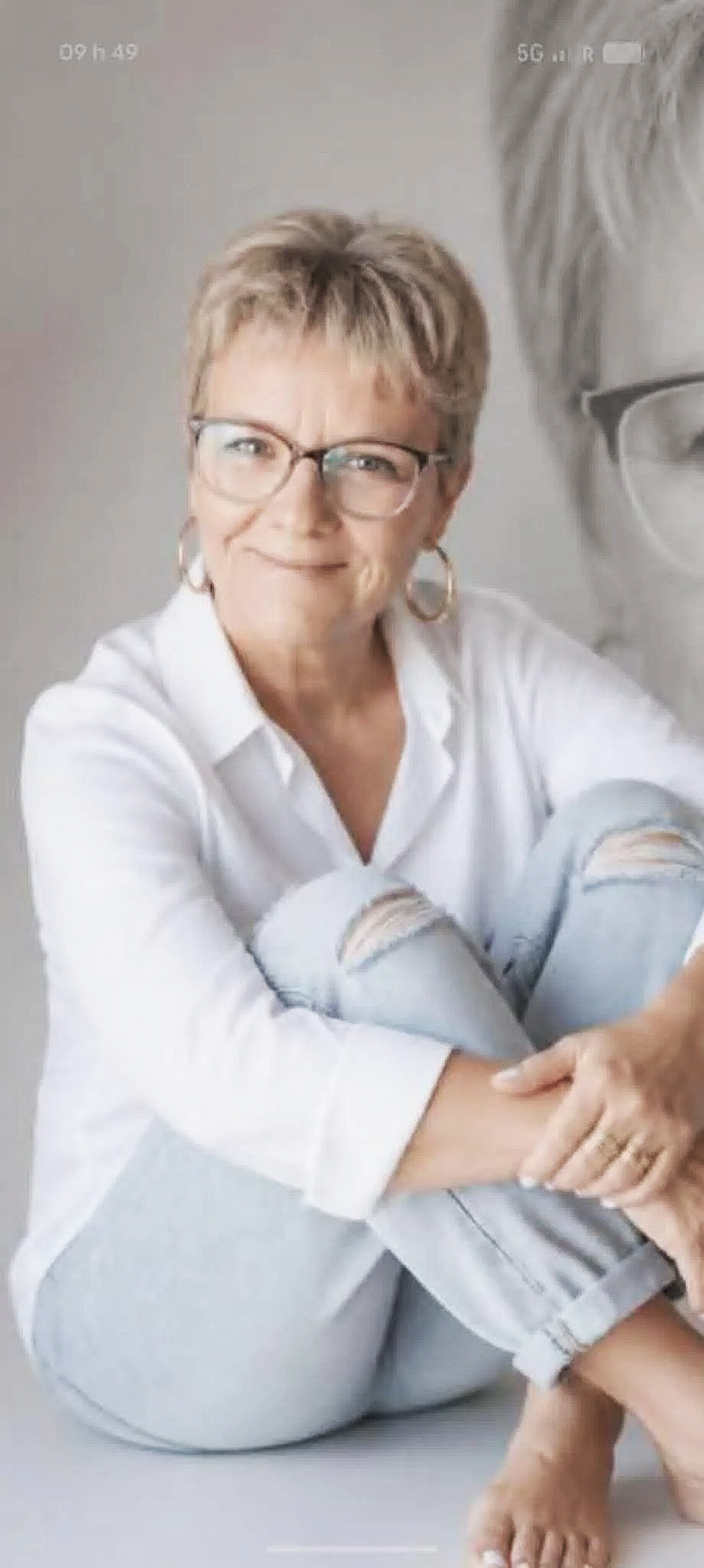 Une femme souriante portant des lunettes, une chemise blanche et des jeans déchirés, assise avec ses bras croisés lors d'une séance photo.