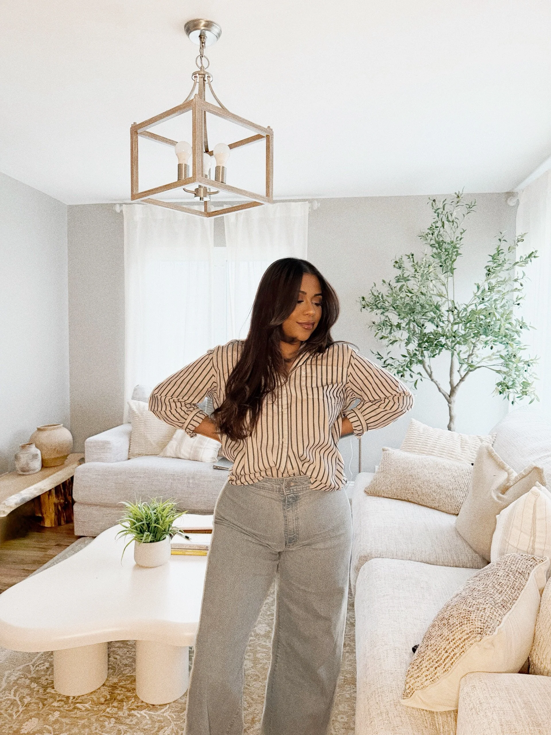 Young woman standing in a cozy, well-decorated living room with her hands on her hips, wearing a striped shirt and gray jeans, with a smile and looking down.