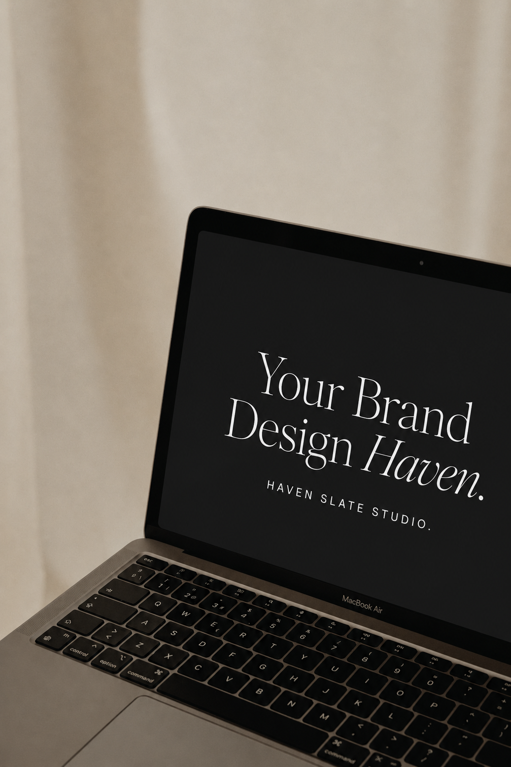 A MacBook Air laptop displaying a black screen with white text that reads "Your Brand Design Haven. HAVEN SLATE STUDIO." The laptop is positioned on a surface against a neutral background.