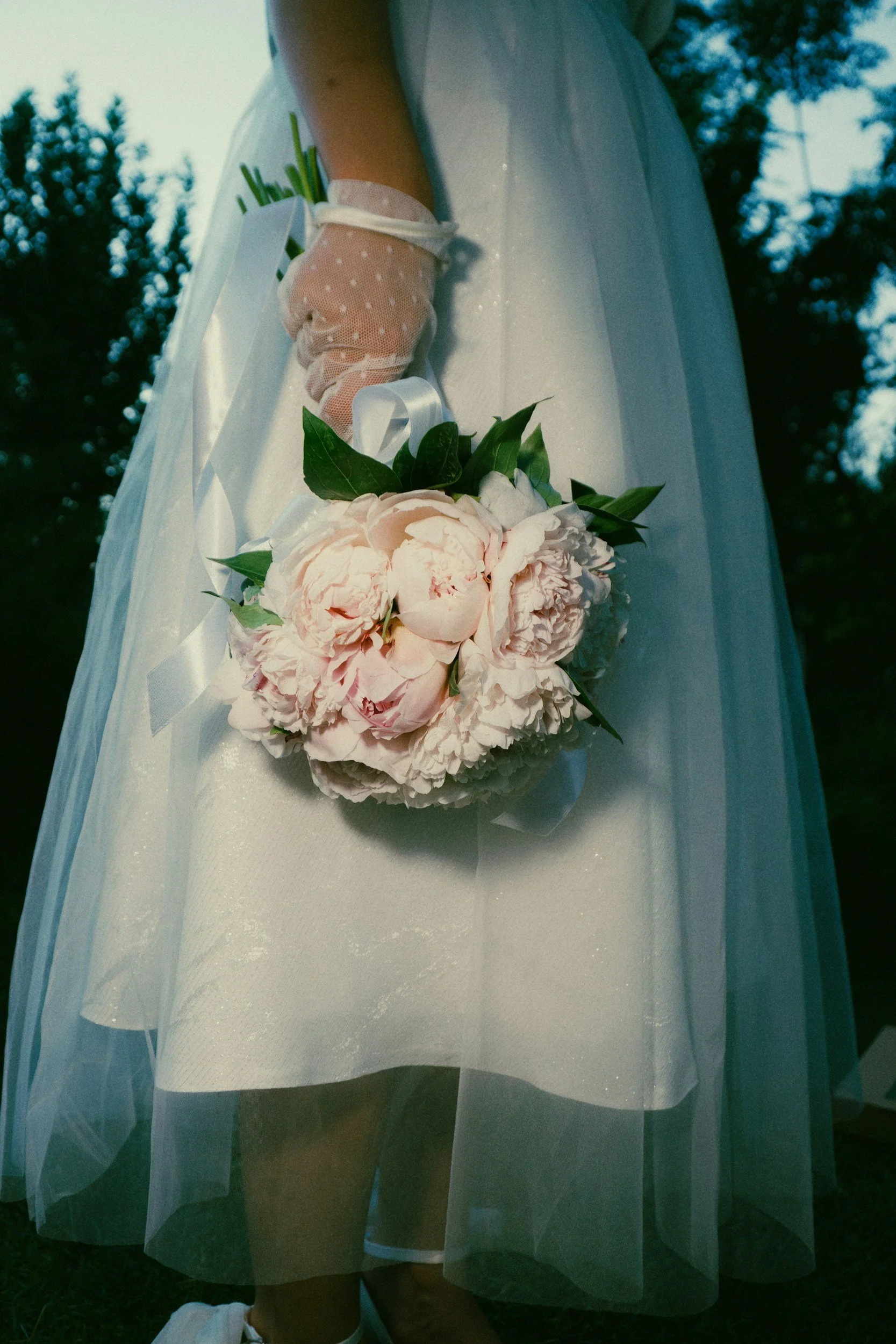 A person wearing a white dress with a tulle overlay, holding a bouquet of pink peonies and greenery, with a garden or outdoor setting in the background.