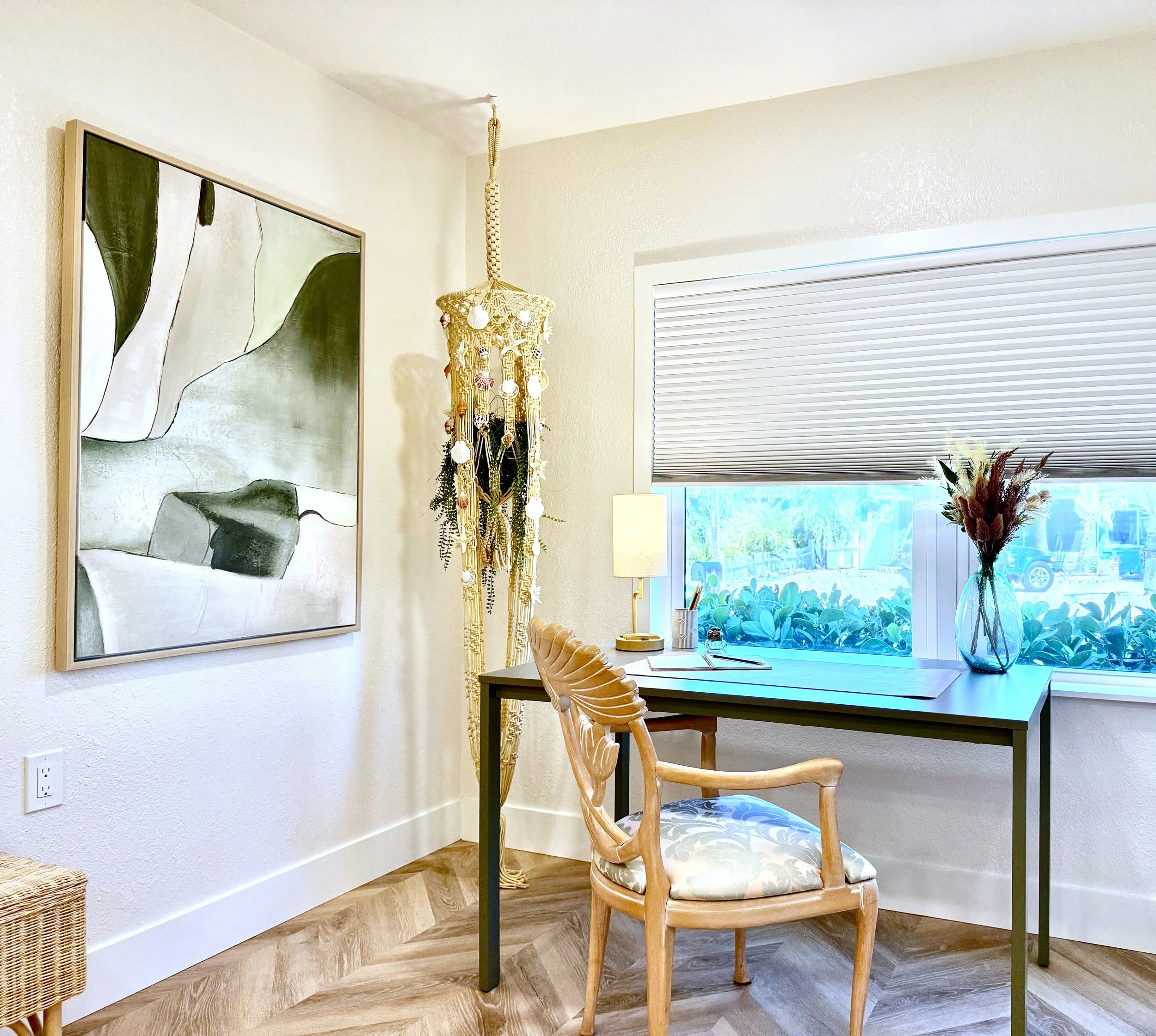 A cozy dining nook with a wooden table and two matching chairs, a large abstract painting on the wall, a macrame hanging decoration, a window with closed blinds, a glass vase with dried flowers, and a table lamp, in a room with wood flooring and white walls.