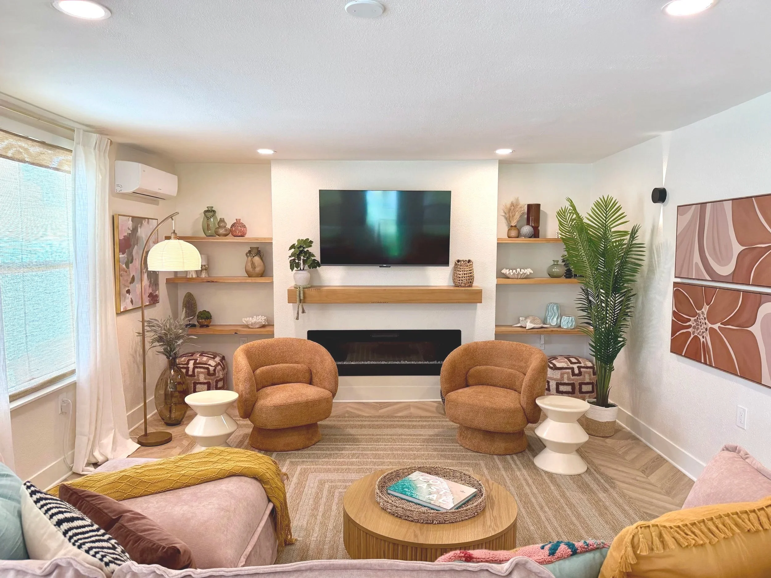 Living room with a mounted flat-screen TV above a wooden fireplace, surrounded by decorative vases, plants, and artwork. Two rounded armchairs face each other in front of a round wooden coffee table, with a large potted plant on the right. There are 
