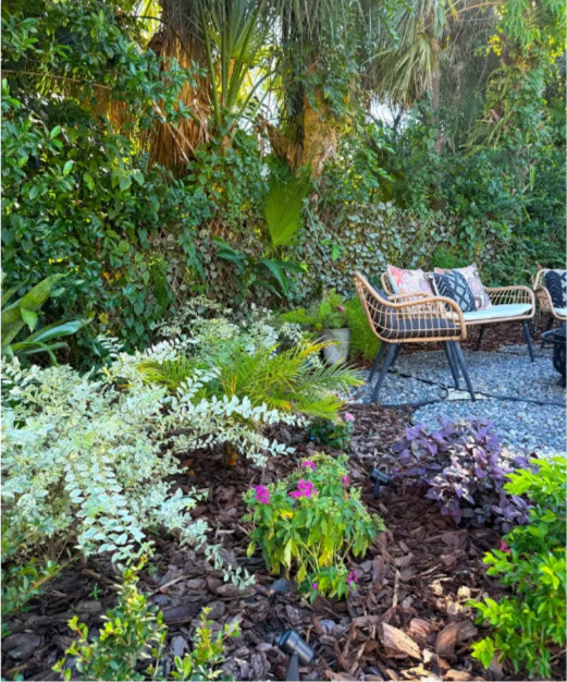 A lush garden with various green plants, flowers, and a seating area with wicker chairs and cushions.