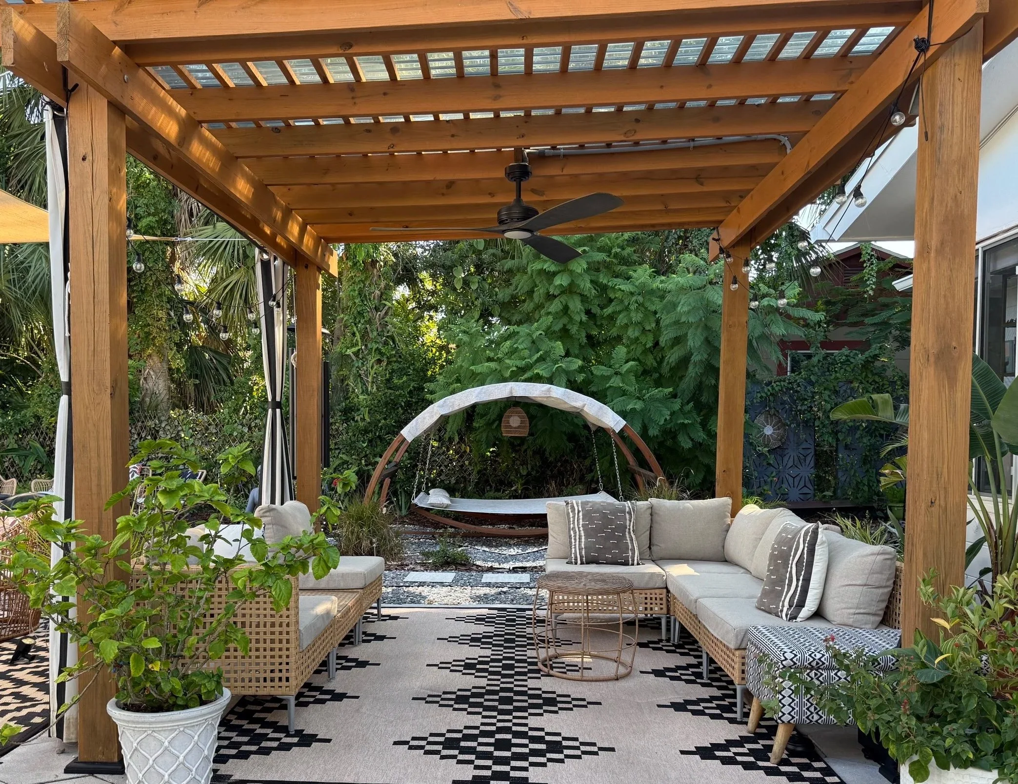 A cozy outdoor patio area with a wooden pergola, ceiling fan, comfortable seating, plants, and a swing in a lush garden setting.
