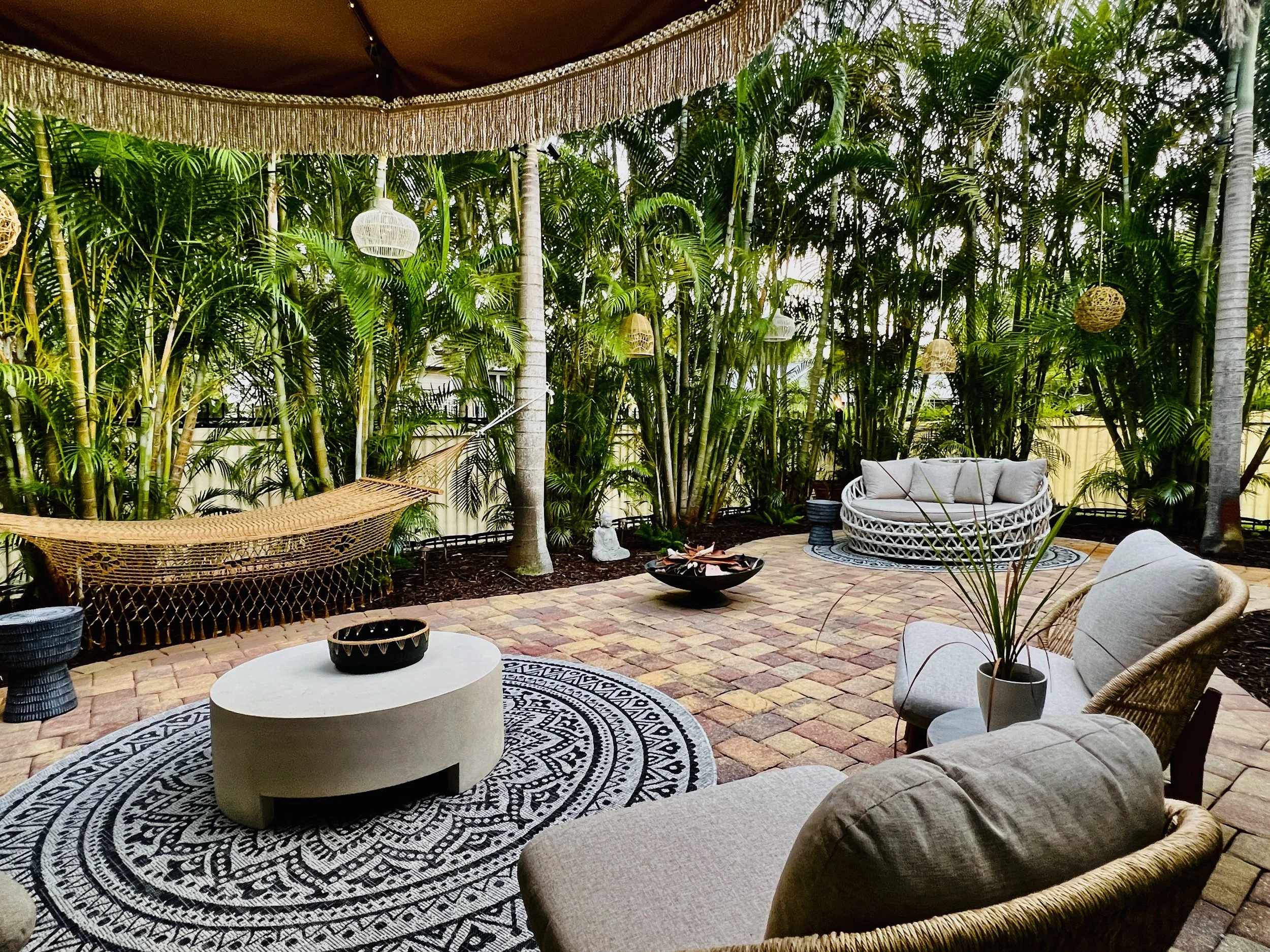 A cozy outdoor patio area surrounded by lush green tropical plants and tall trees. The space features wicker and upholstered furniture, decorative hanging lanterns, a colorful area rug, and a brick paving base.