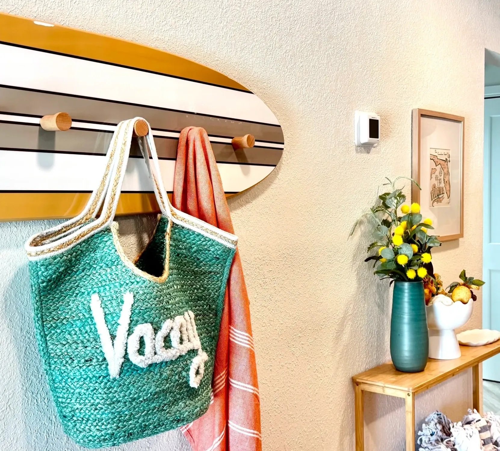 Decorative wall with hanging bag and towels, framed artwork, a tall teal vase with yellow flowers, and small decorative objects on a wooden table.