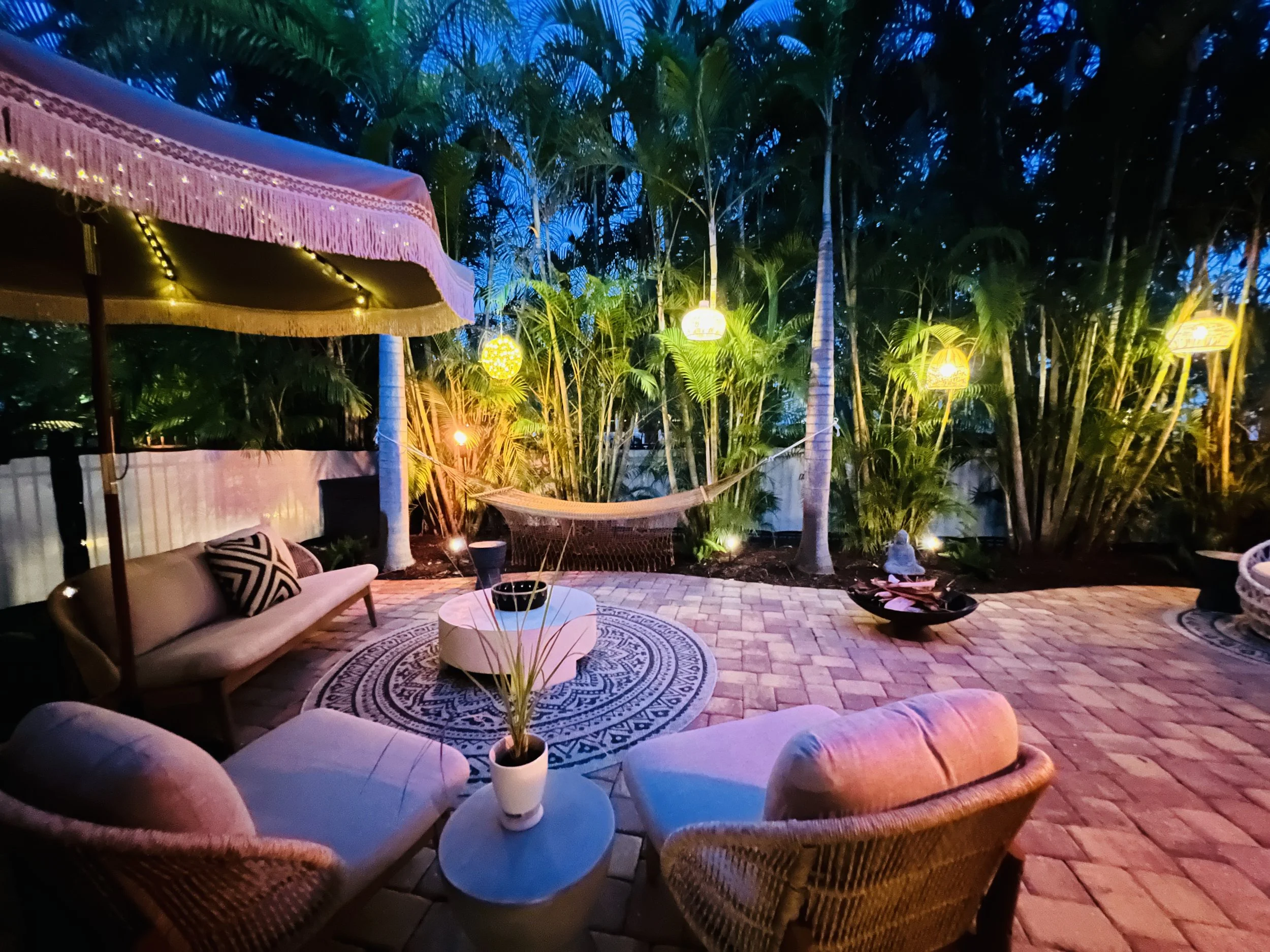 A cozy outdoor patio area at dusk, featuring a sofa, wicker chairs, a small round table, potted plants, and hanging lanterns among tall palm trees, with string lights adding warm ambiance.